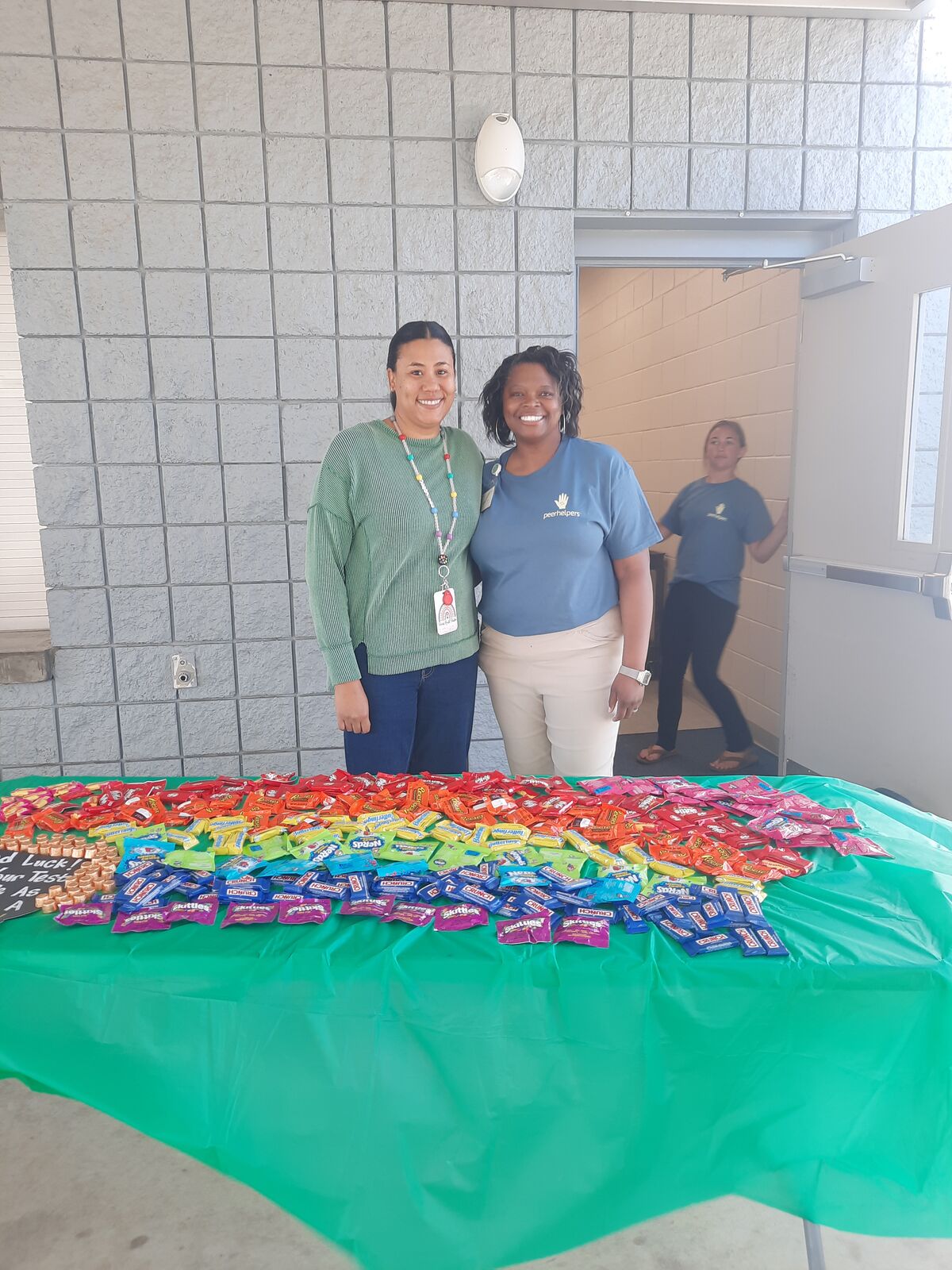 Two smiling women stand behind a table covered with a green cloth, laden with a colorful assortment of candy.