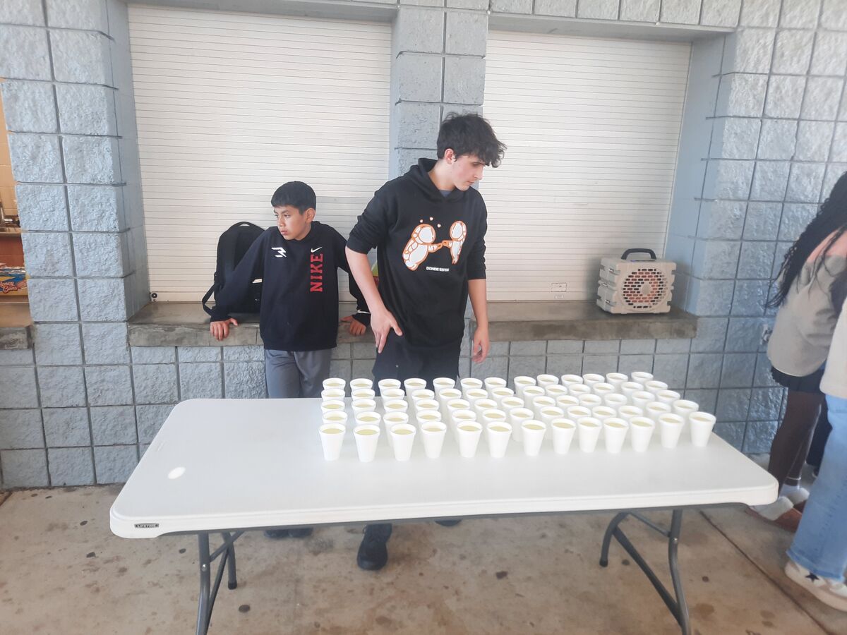 Two young people stand near a table laden with many white cups filled with a light-colored liquid.