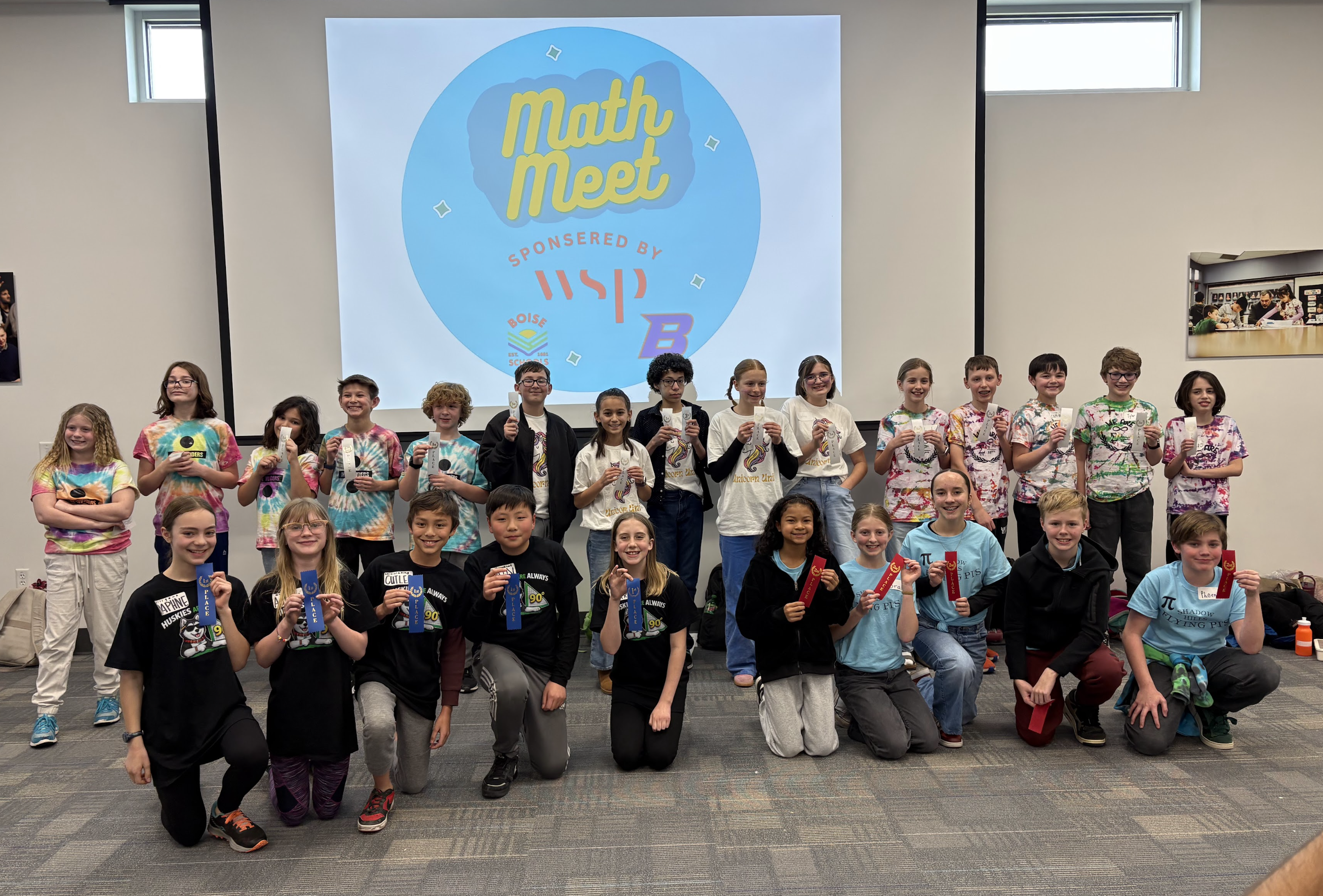 A group of smiling students, some wearing tie-dye shirts and others black t-shirts, pose for a photo holding award ribbons in front of a "Math Meet" banner.