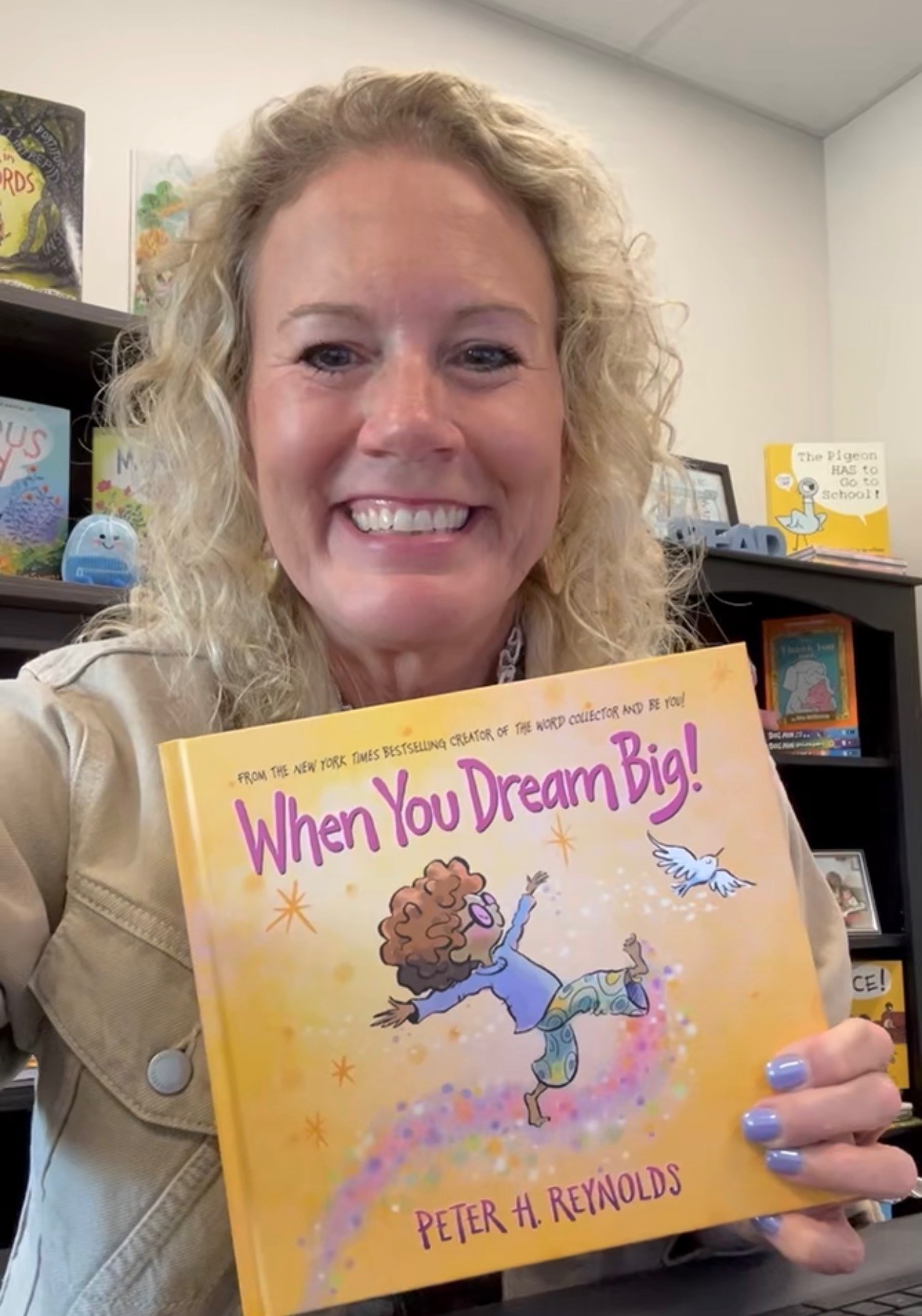 A smiling woman holds up a children's book titled 'When You Dream Big!' by Peter H. Reynolds.