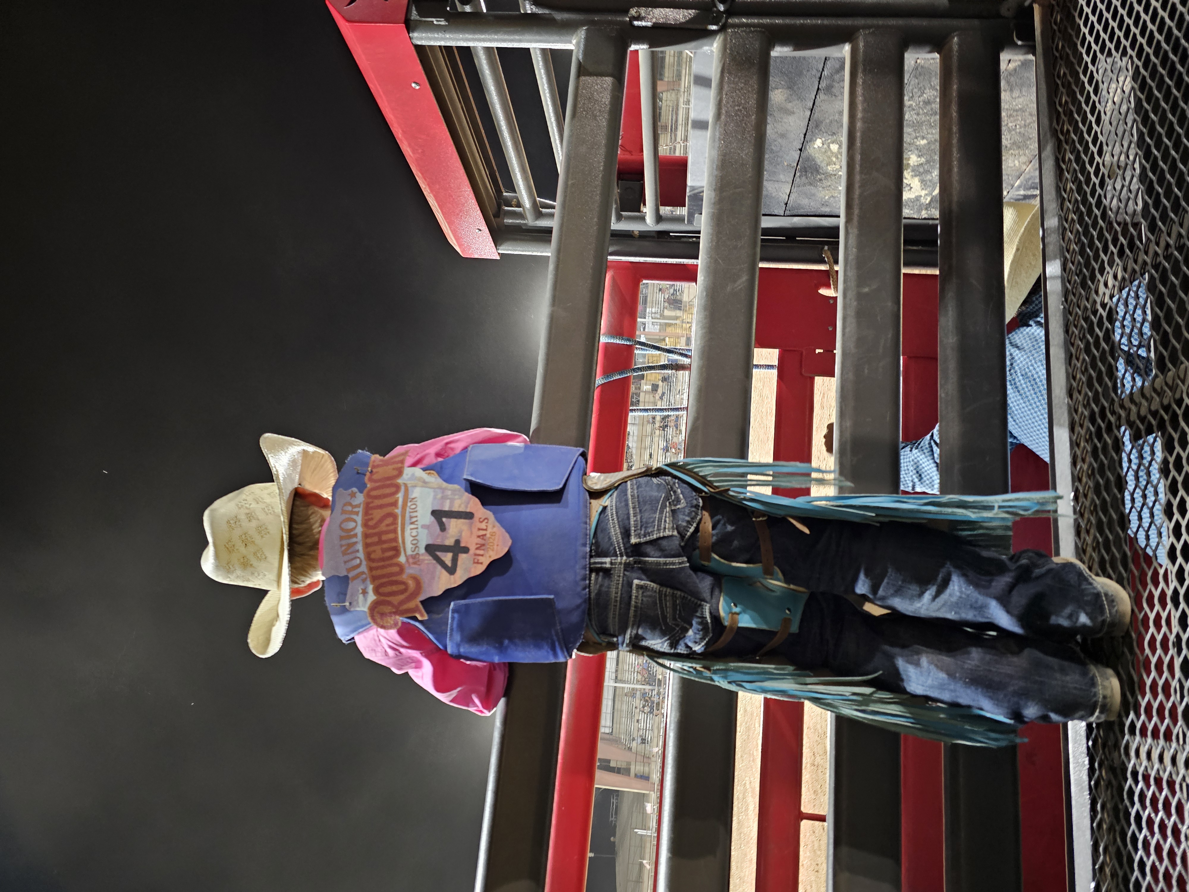 young boy standing on a bucking chute wearing a cowboy hat, chaps and number