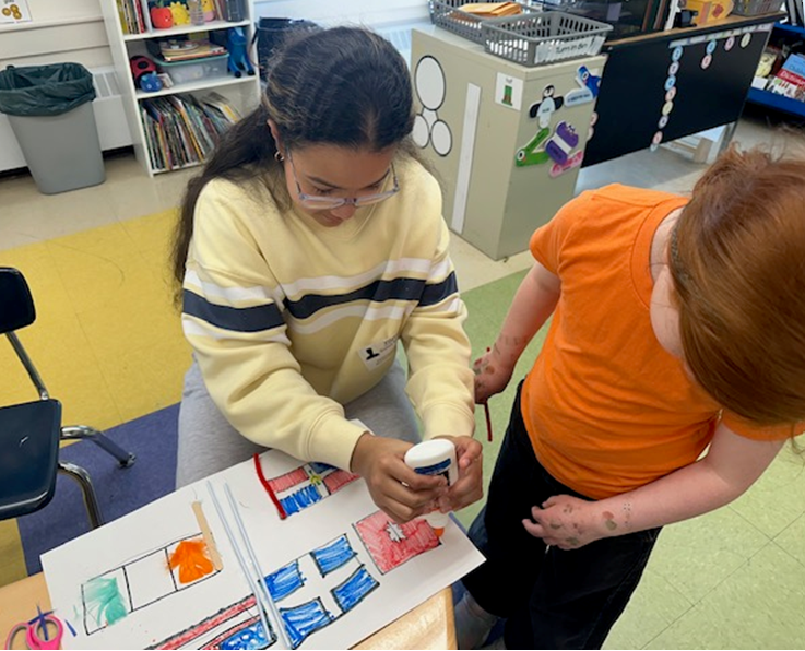 A young girl in a yellow sweater and a younger child in an orange shirt work together on an art project at a table.