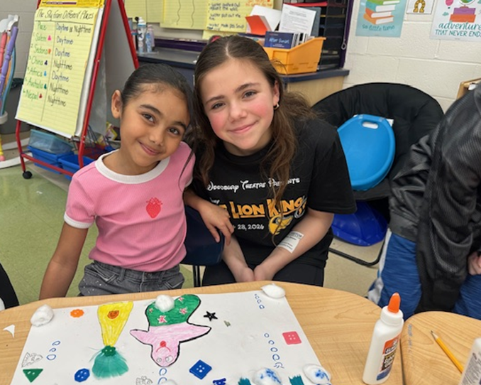 Two young girls smile at the camera in a classroom, with a colorful art project on the table.