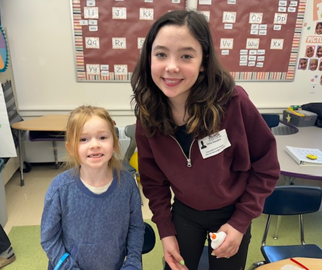Two smiling girls stand in a classroom with alphabet charts on the wall.