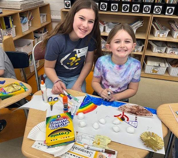 Two smiling girls work on an art project at a classroom desk, surrounded by art supplies.