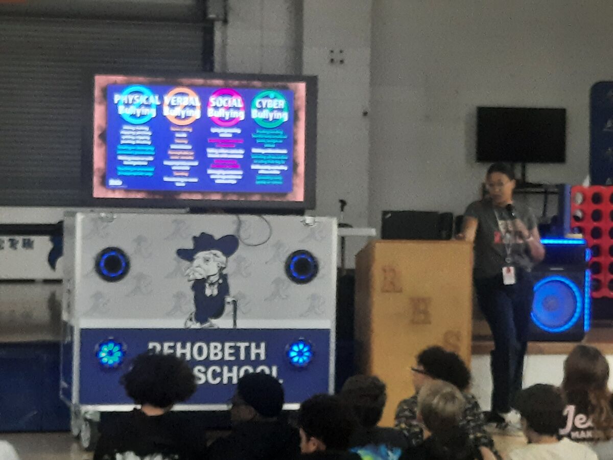 A speaker presents information about physical, verbal, social, and cyber bullying to an audience in a school setting.