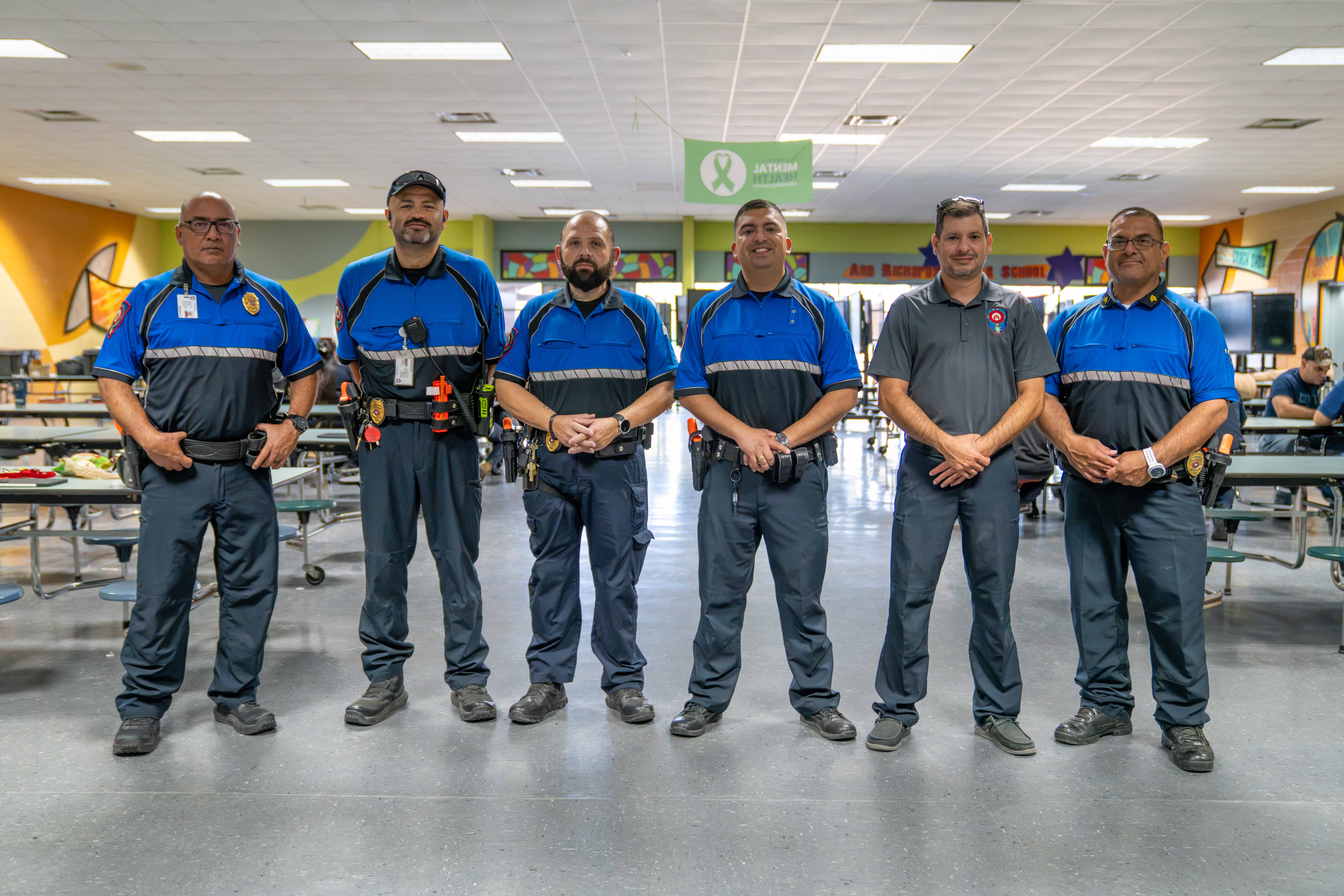 Six security personnel stand in a line inside a school cafeteria, wearing matching uniforms.