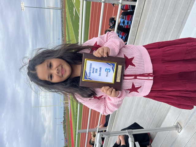 A smiling child holds an award plaque while standing on stadium bleachers with a track and field in the background.