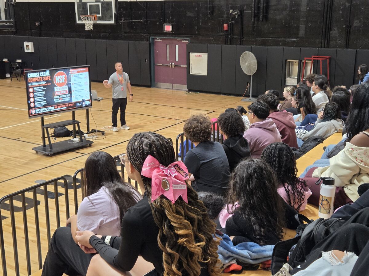 A speaker presents information on a screen to a group of students seated in a school gymnasium.
