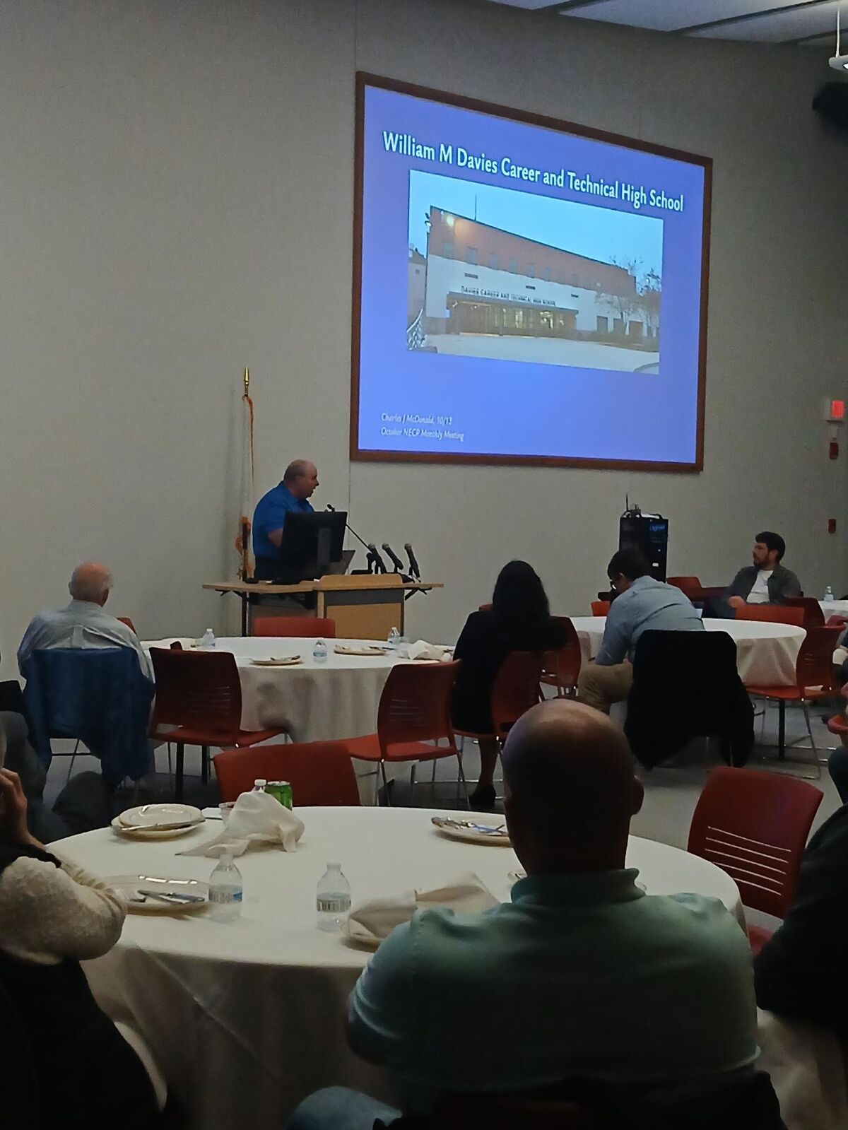 A speaker presents at a podium in a conference room with seated attendees at round tables and a large projection screen displaying a school.