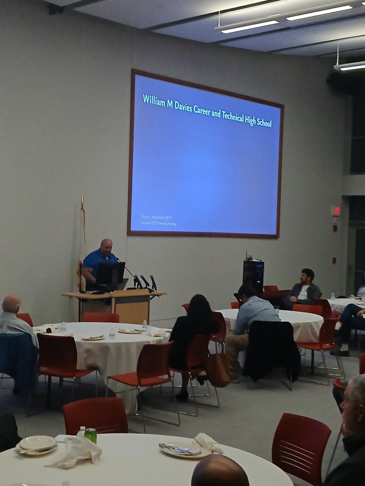 A speaker stands at a podium in a large hall, presenting in front of a projection screen that reads "William M Davies Career and Technical High School."