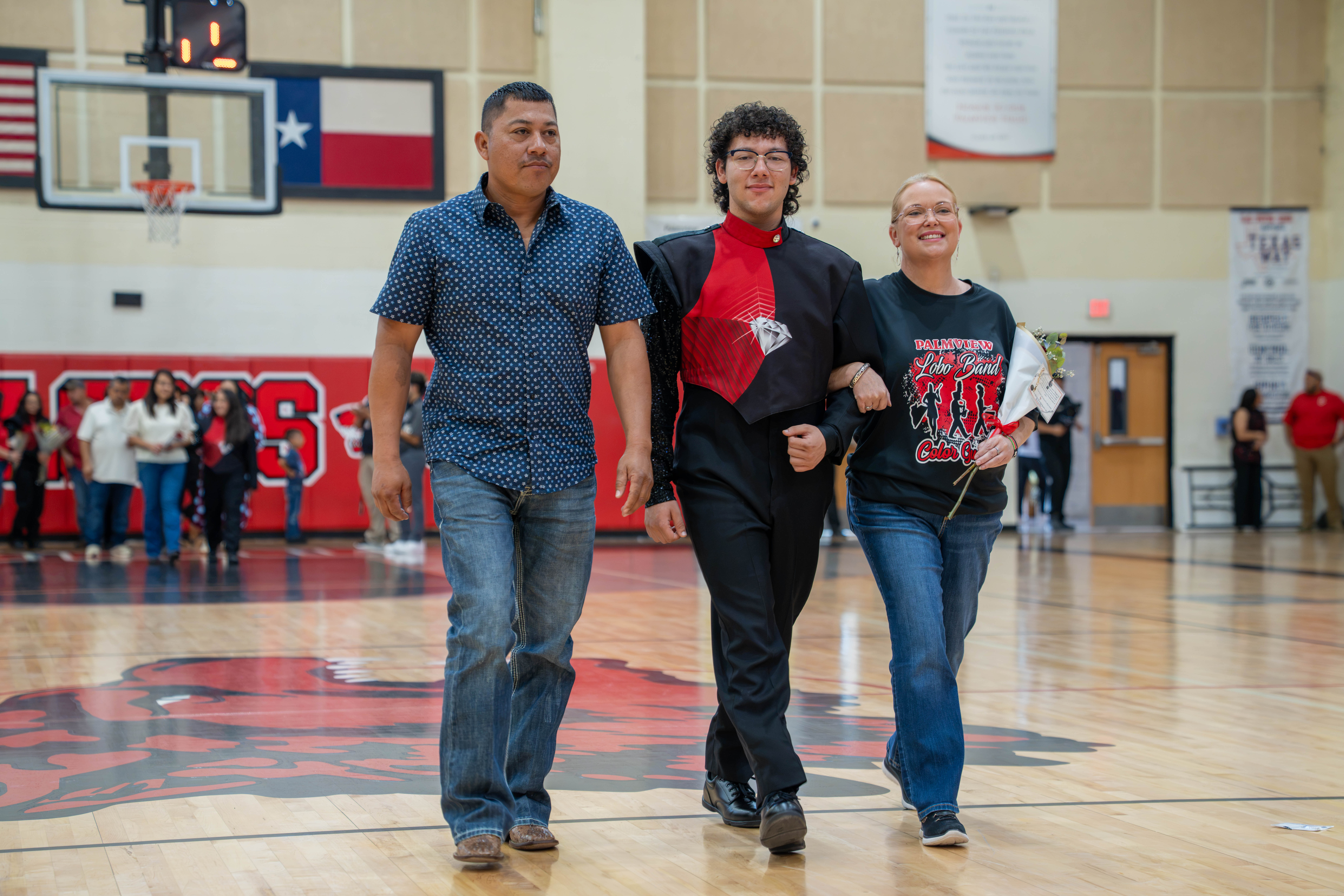 A young person in a marching band uniform is escorted by two adults on a basketball court.