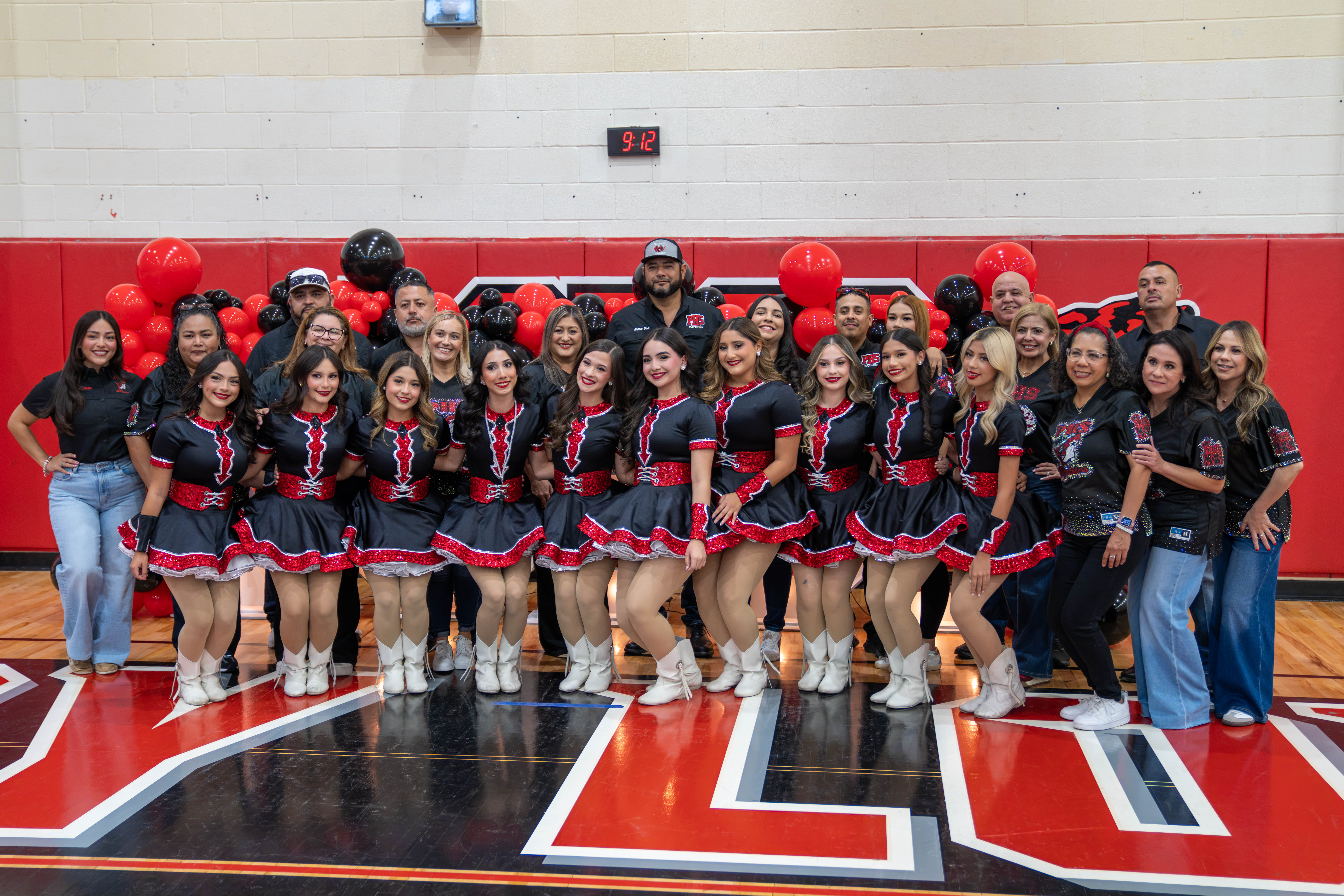 A group of dancers in matching black and red outfits pose for a photo with adults in a gymnasium.