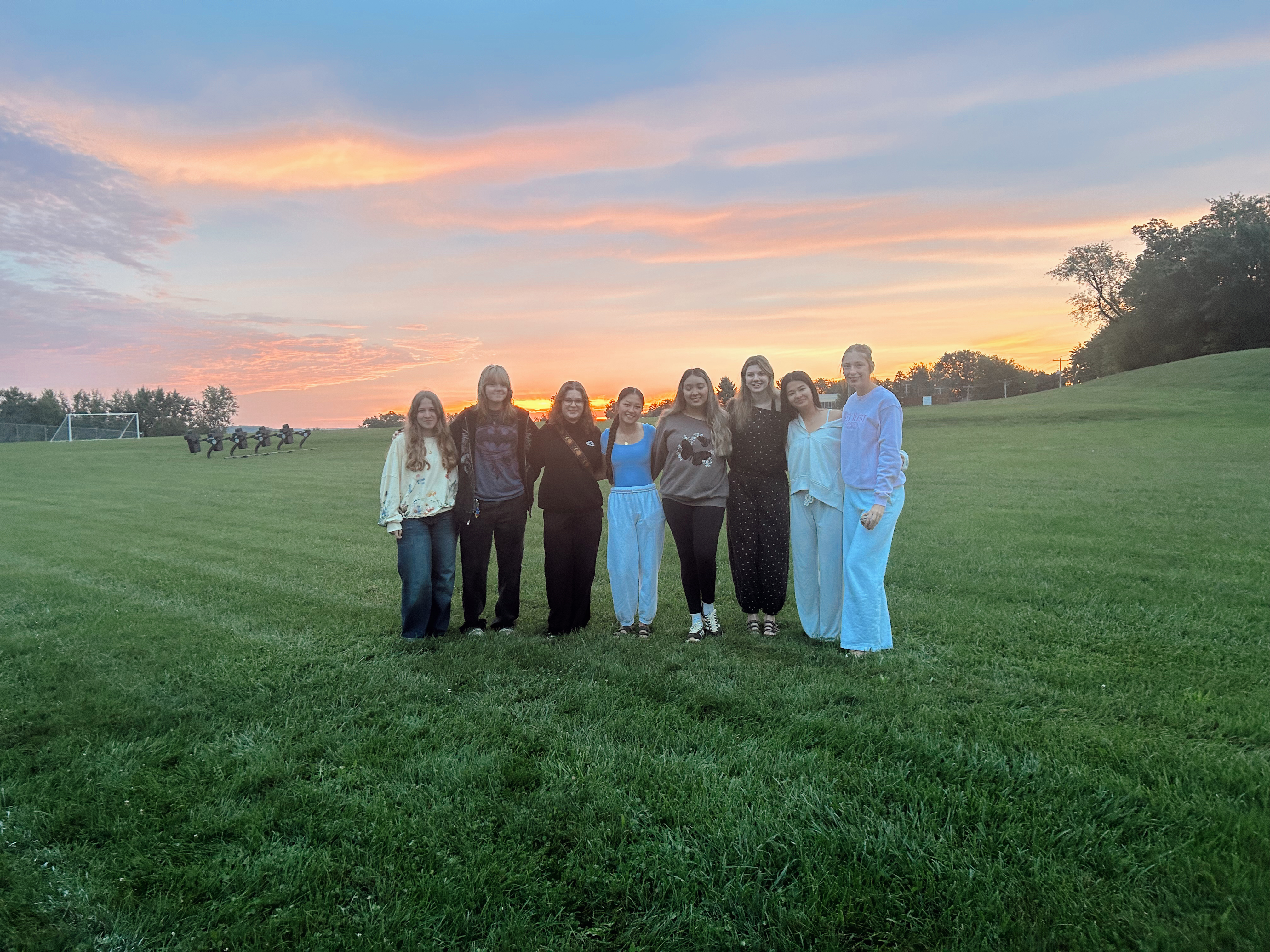 A group of eight friends stand in a grassy field during a vibrant sunrise.