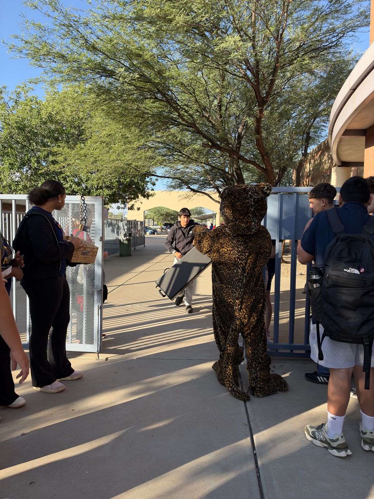 A person in a jaguar mascot costume stands outdoors near a gate as students walk by on a sunny day.