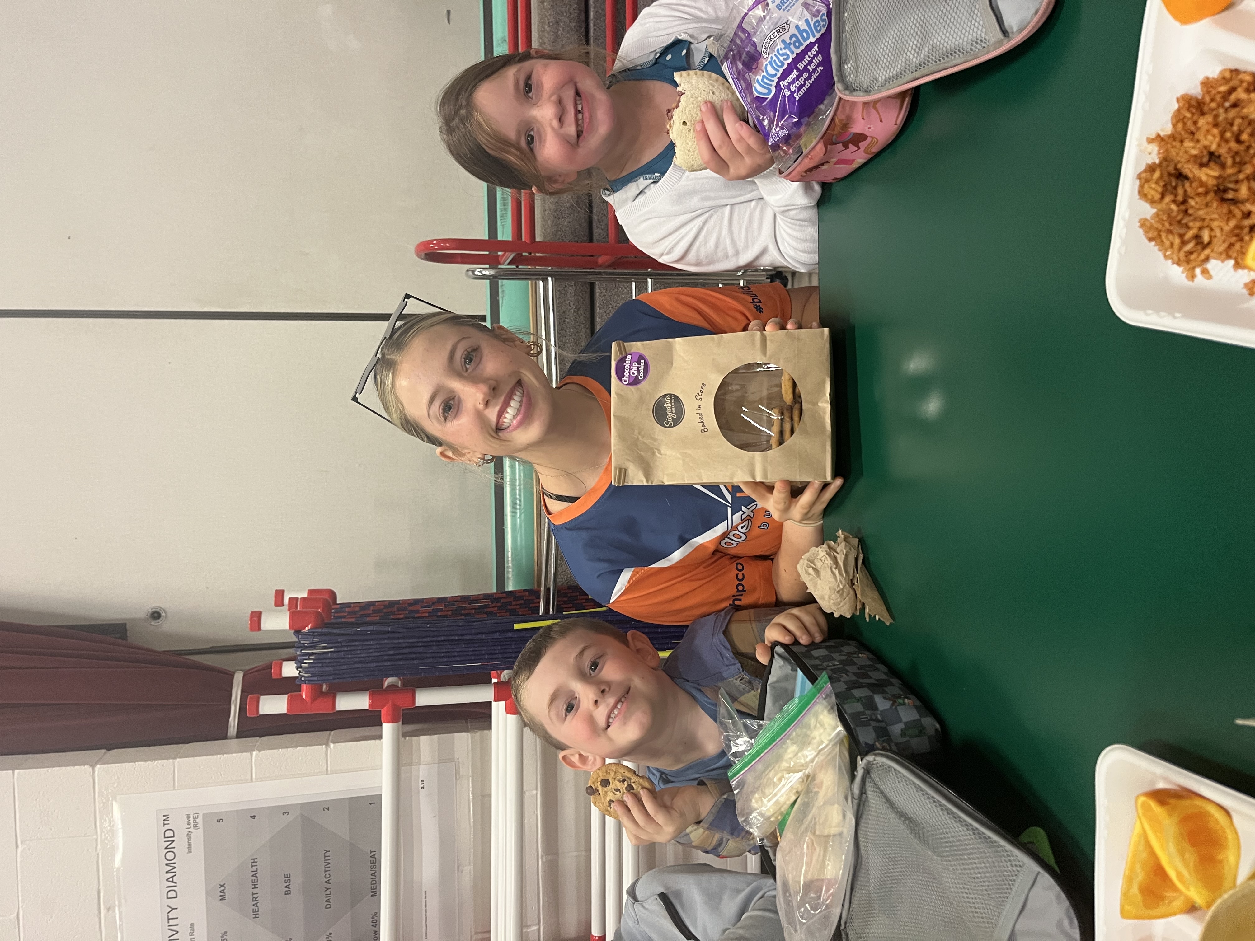 A smiling woman and two children sit at a green table, holding snacks and lunch items.