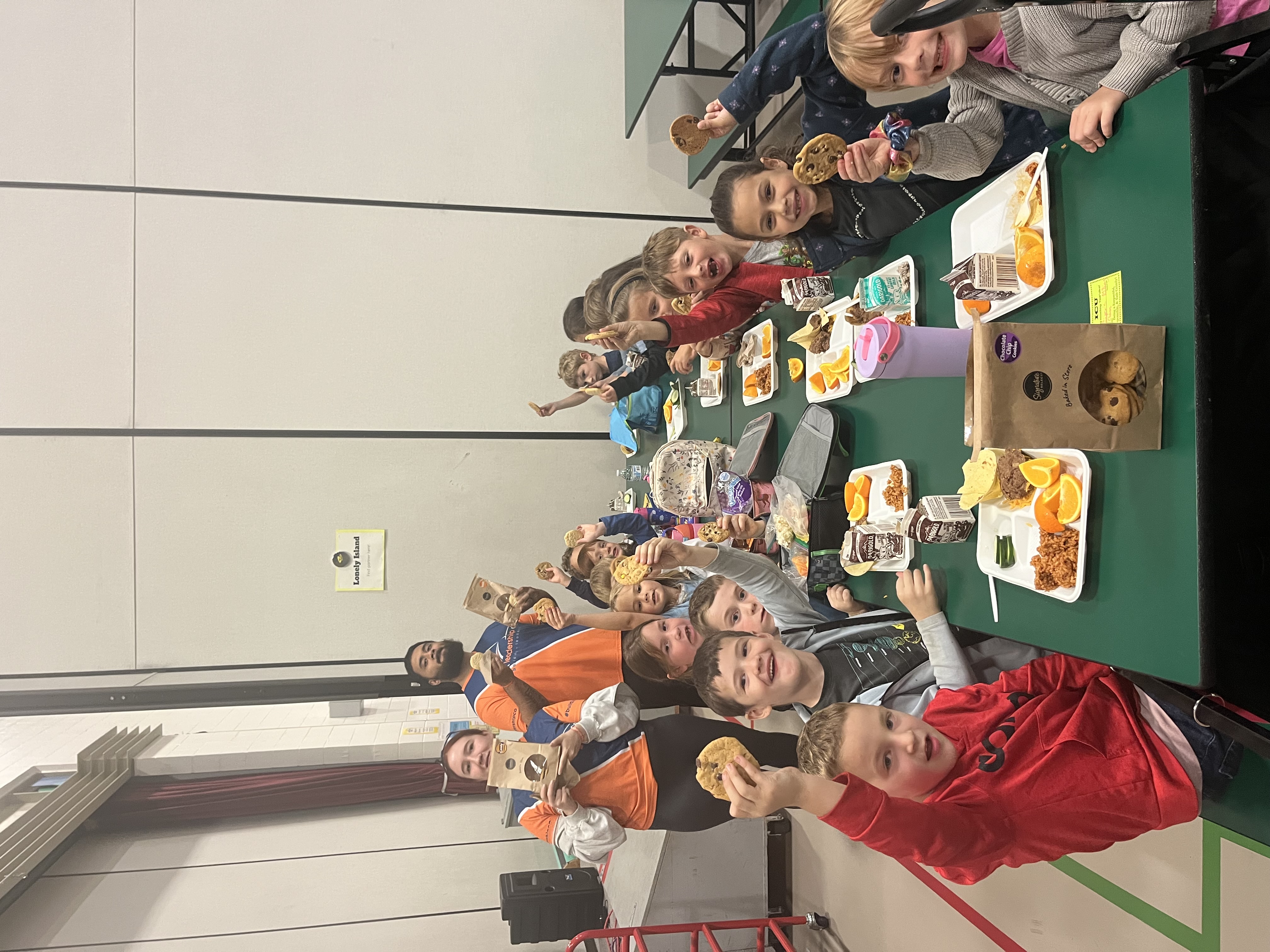 A group of children and adults sit along a long green table, smiling and holding up cookies.