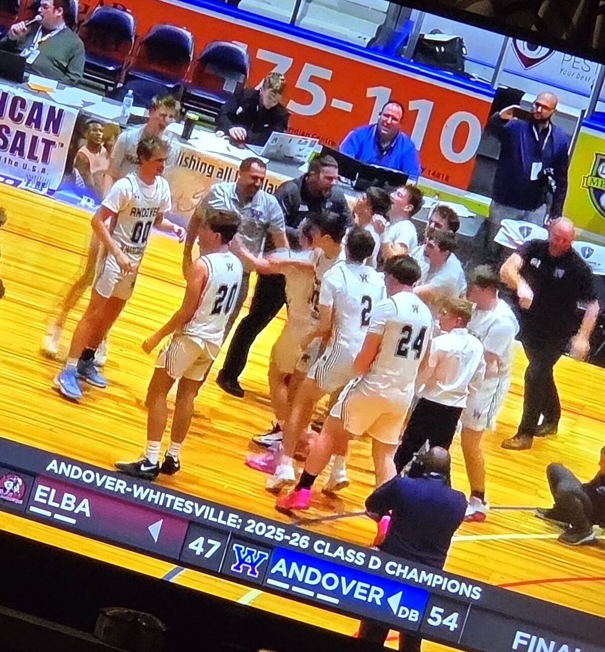 A basketball team celebrates on the court after winning the 2025-26 Class D Championship, with the score showing Andover 54, Elba 47.