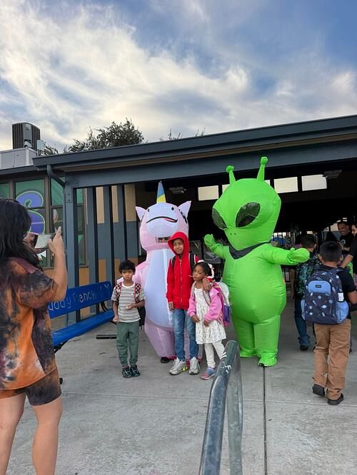 Three children pose with people in inflatable unicorn and alien costumes outside a school.