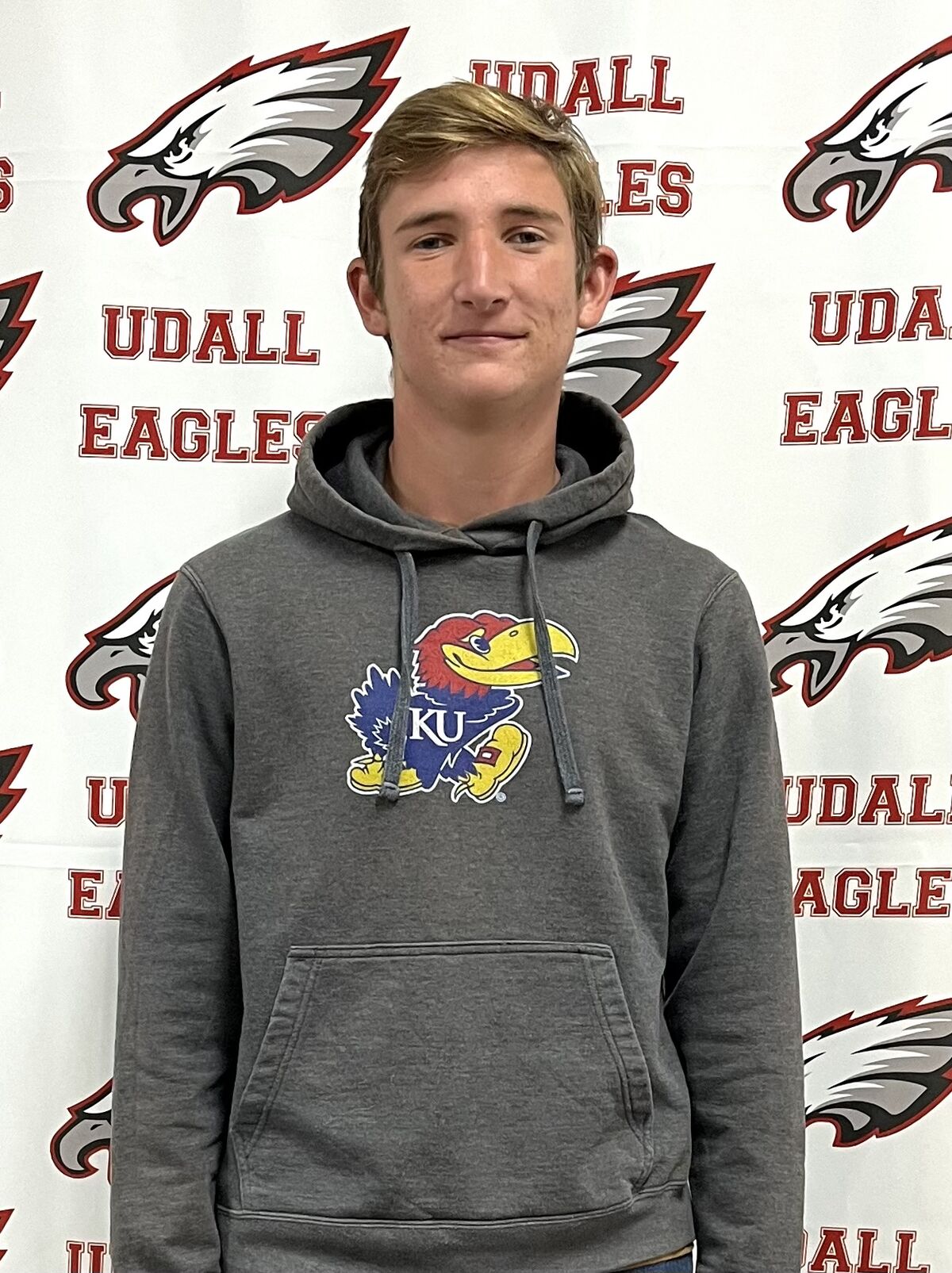 A young person with short blond hair wears a gray hoodie with a KU logo, standing in front of a backdrop featuring 'UDALL EAGLES' text and eagle graphics.