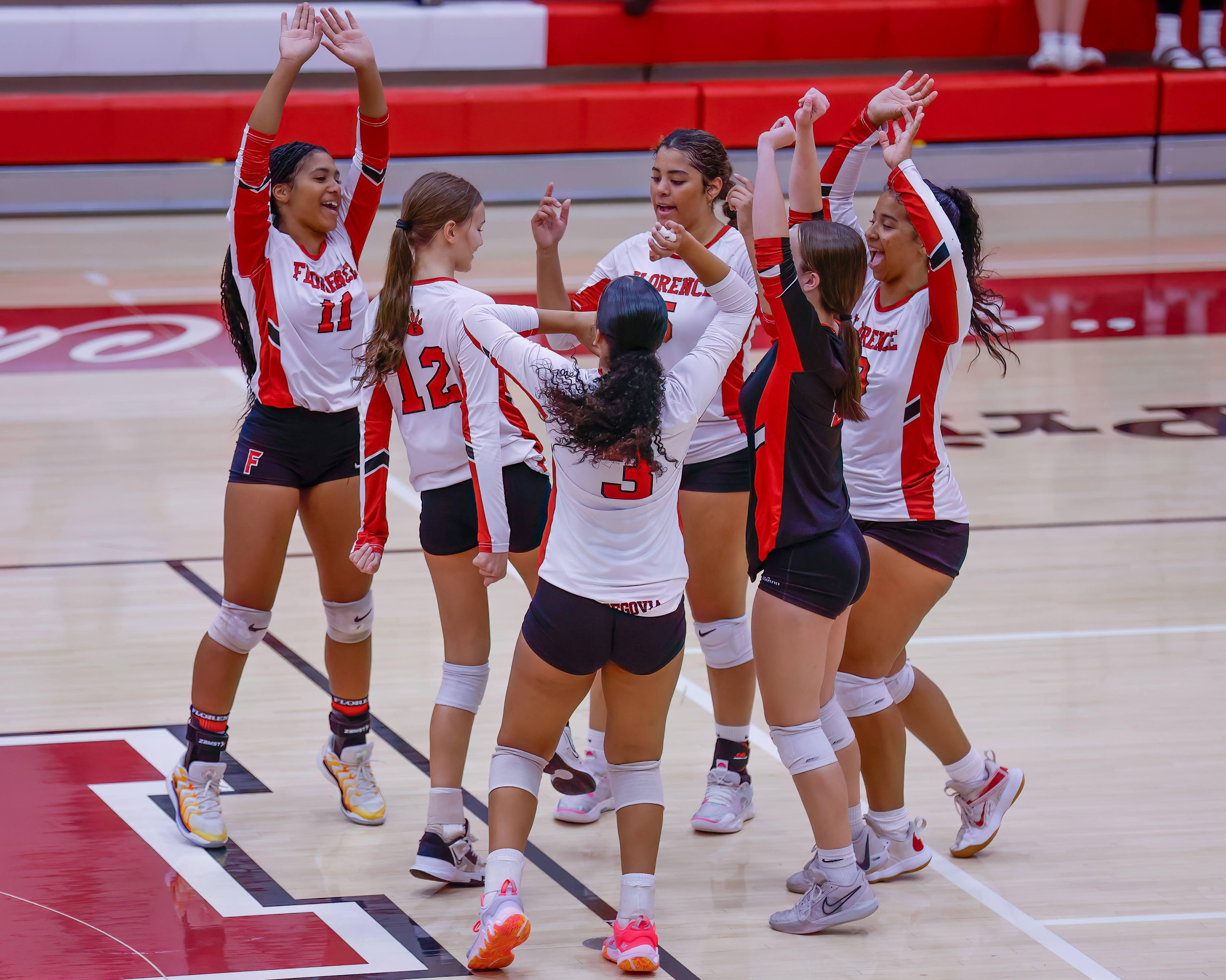 A group of young women in volleyball uniforms celebrate together on a court.