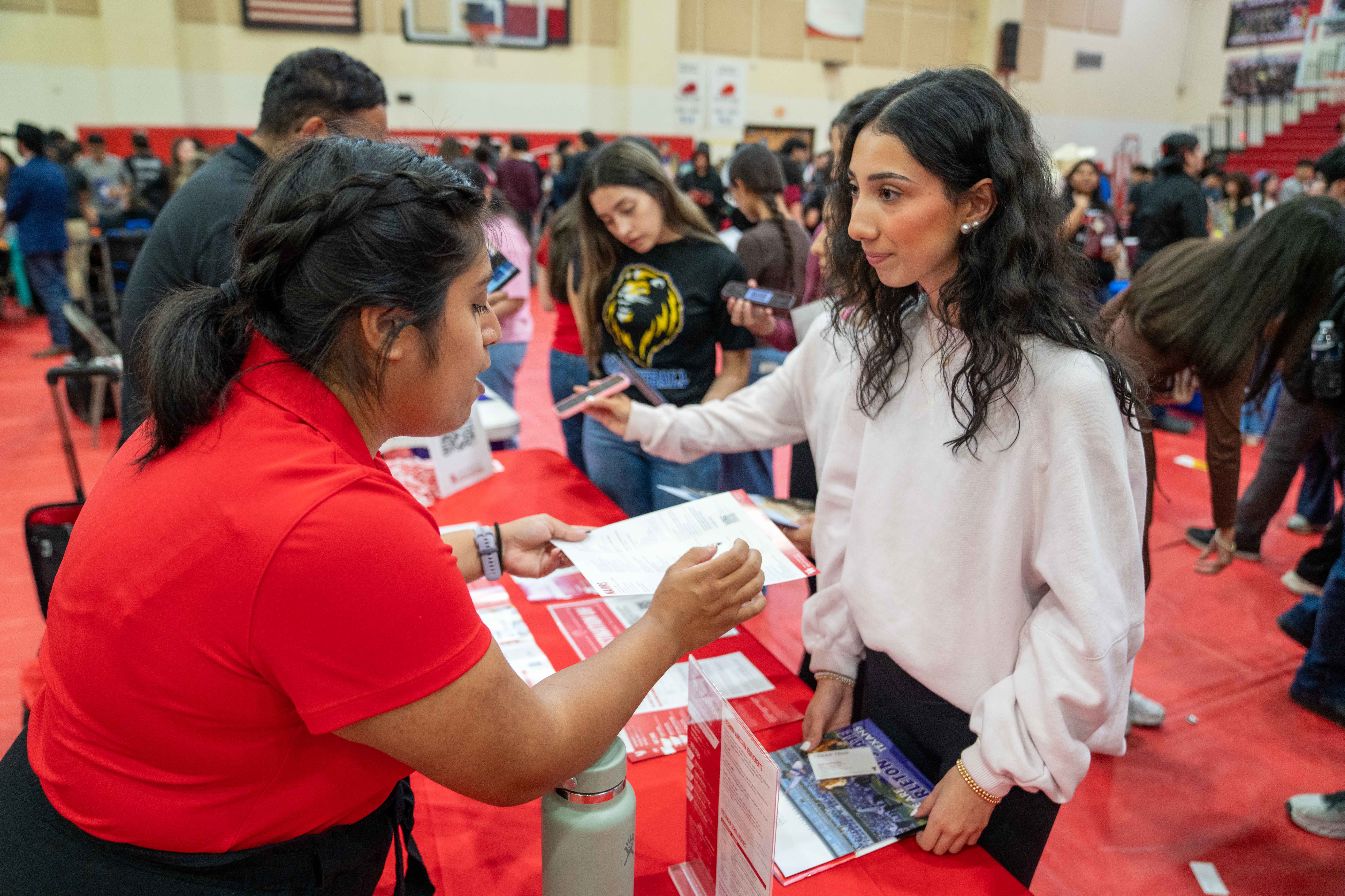 A student in a white sweater talks to a representative at a table during a crowded school career or college fair.