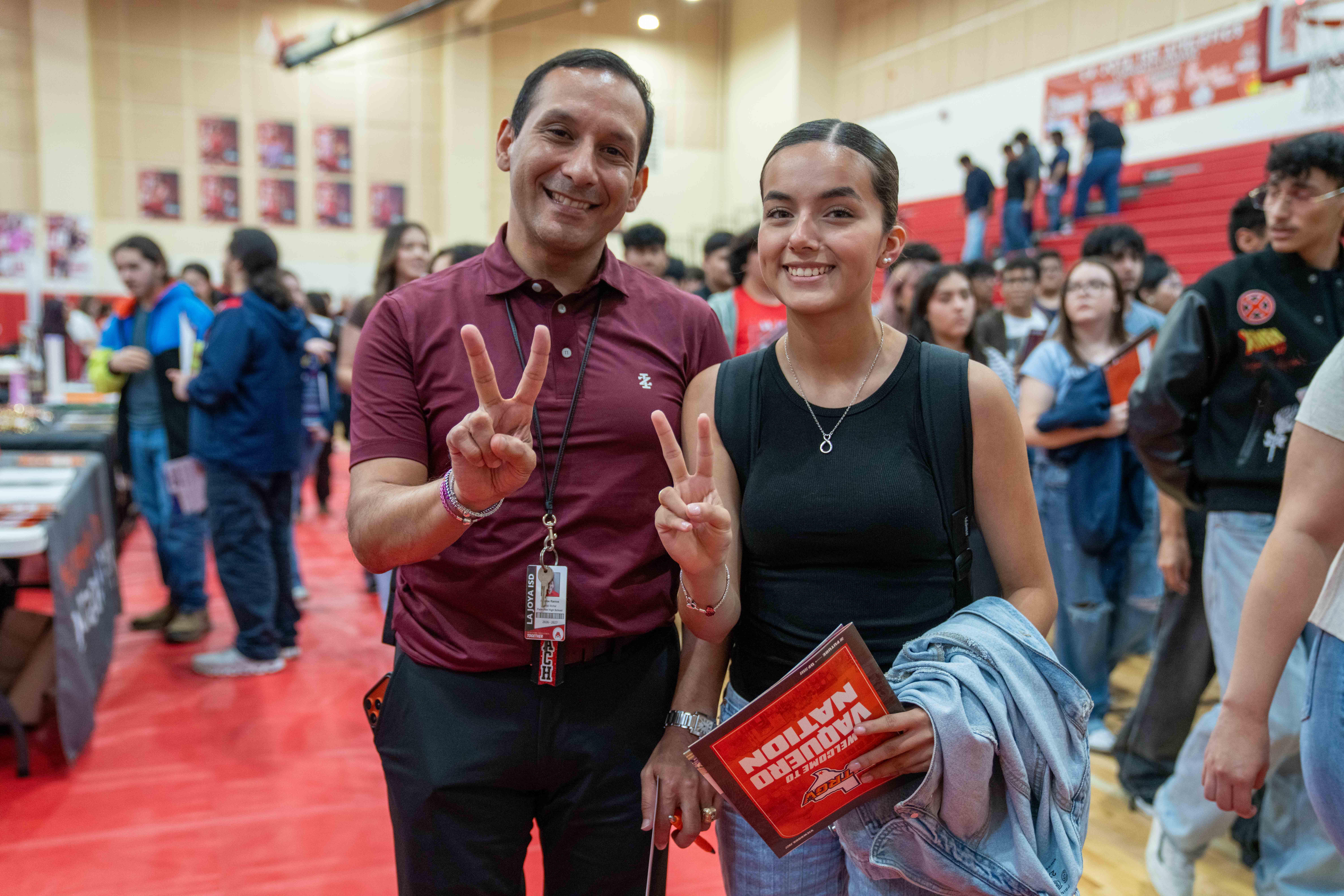 A smiling man and a young woman pose together inside a gymnasium, both holding up peace signs for the camera.