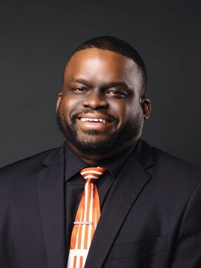 A man in a black suit and an orange and white striped tie smiles warmly.