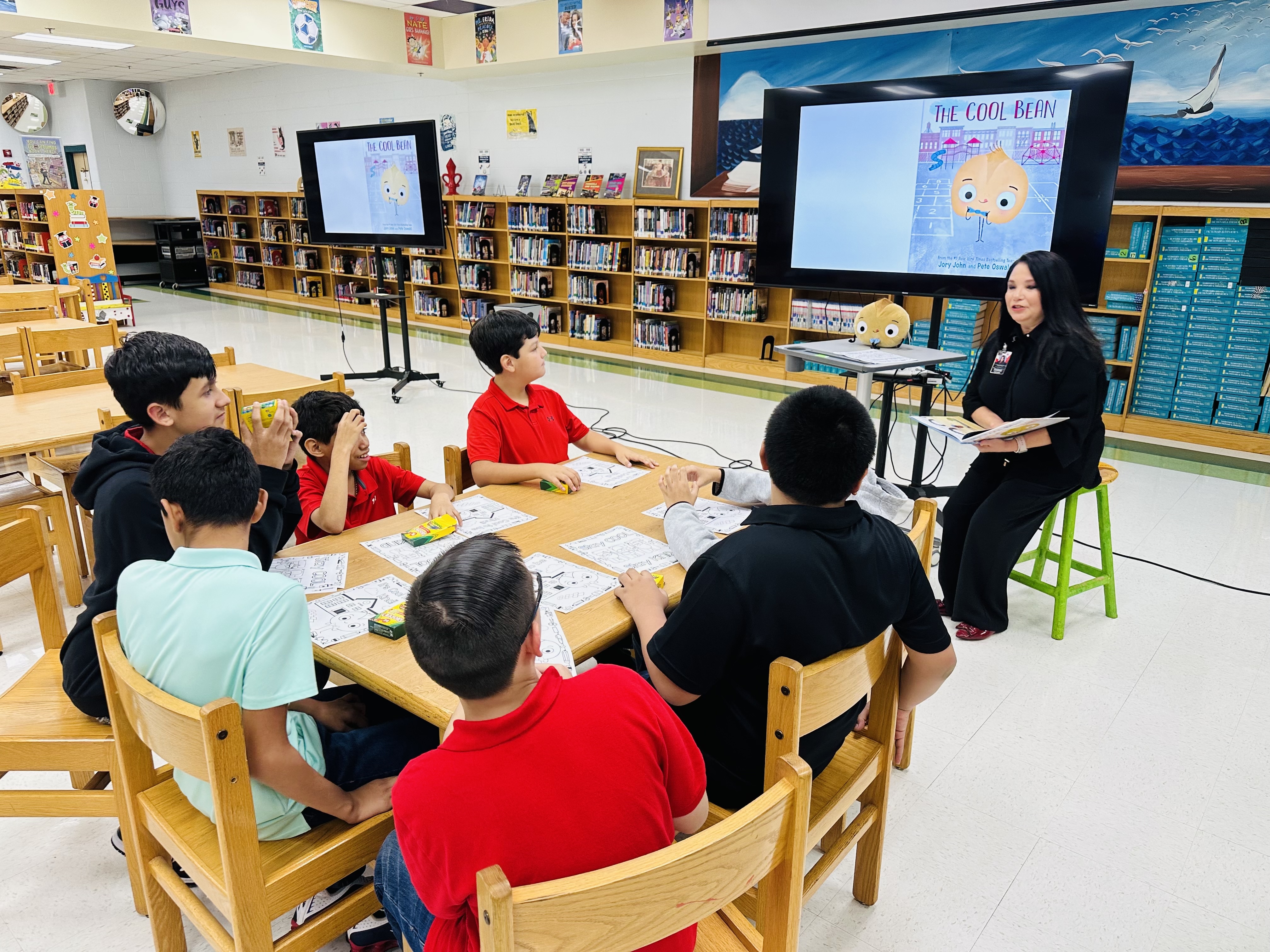 A teacher reads "The Cool Bean" to a group of attentive students seated around a wooden table in a school library.