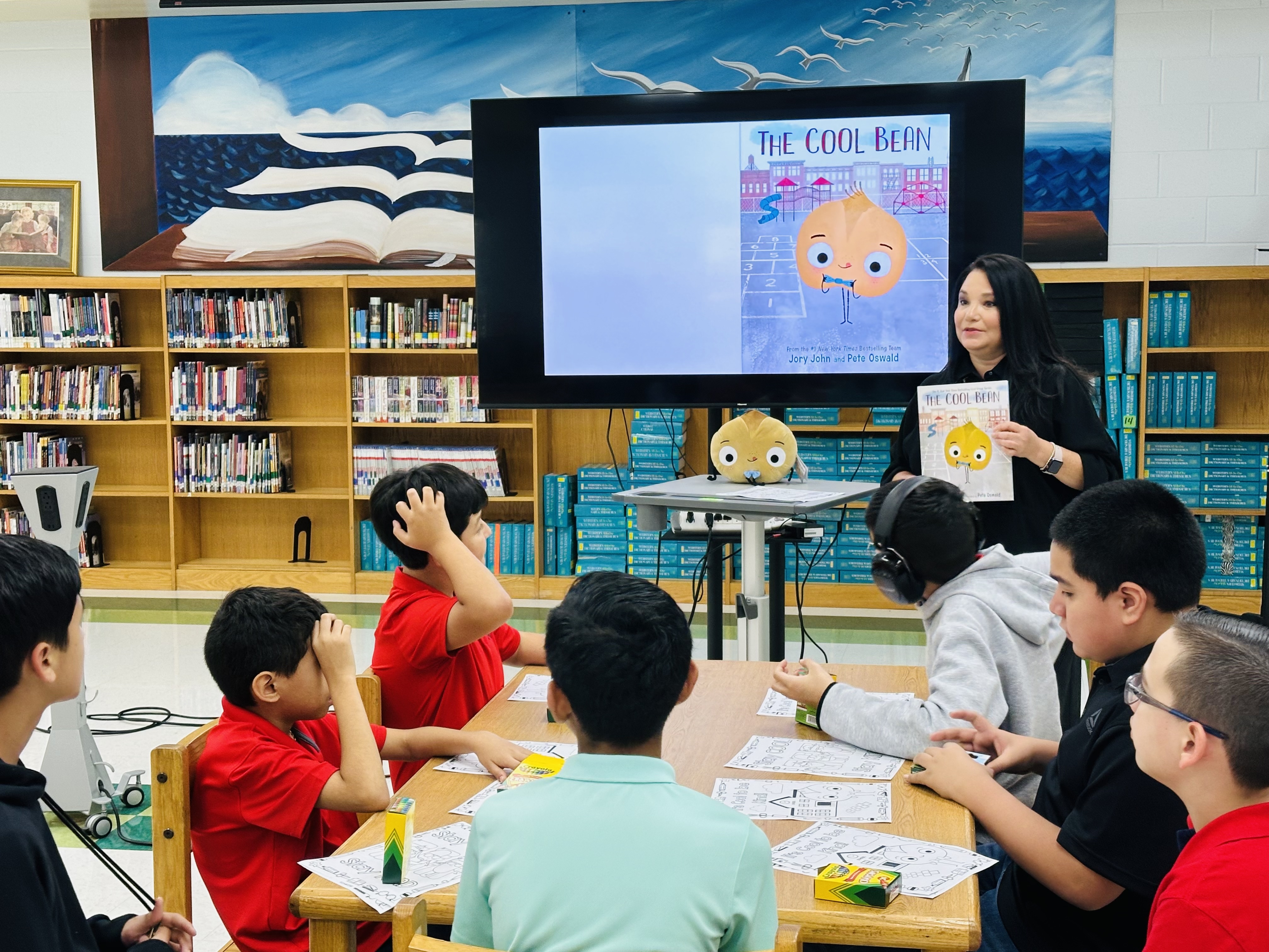 A teacher reads "The Cool Bean" to a group of attentive students in a library.