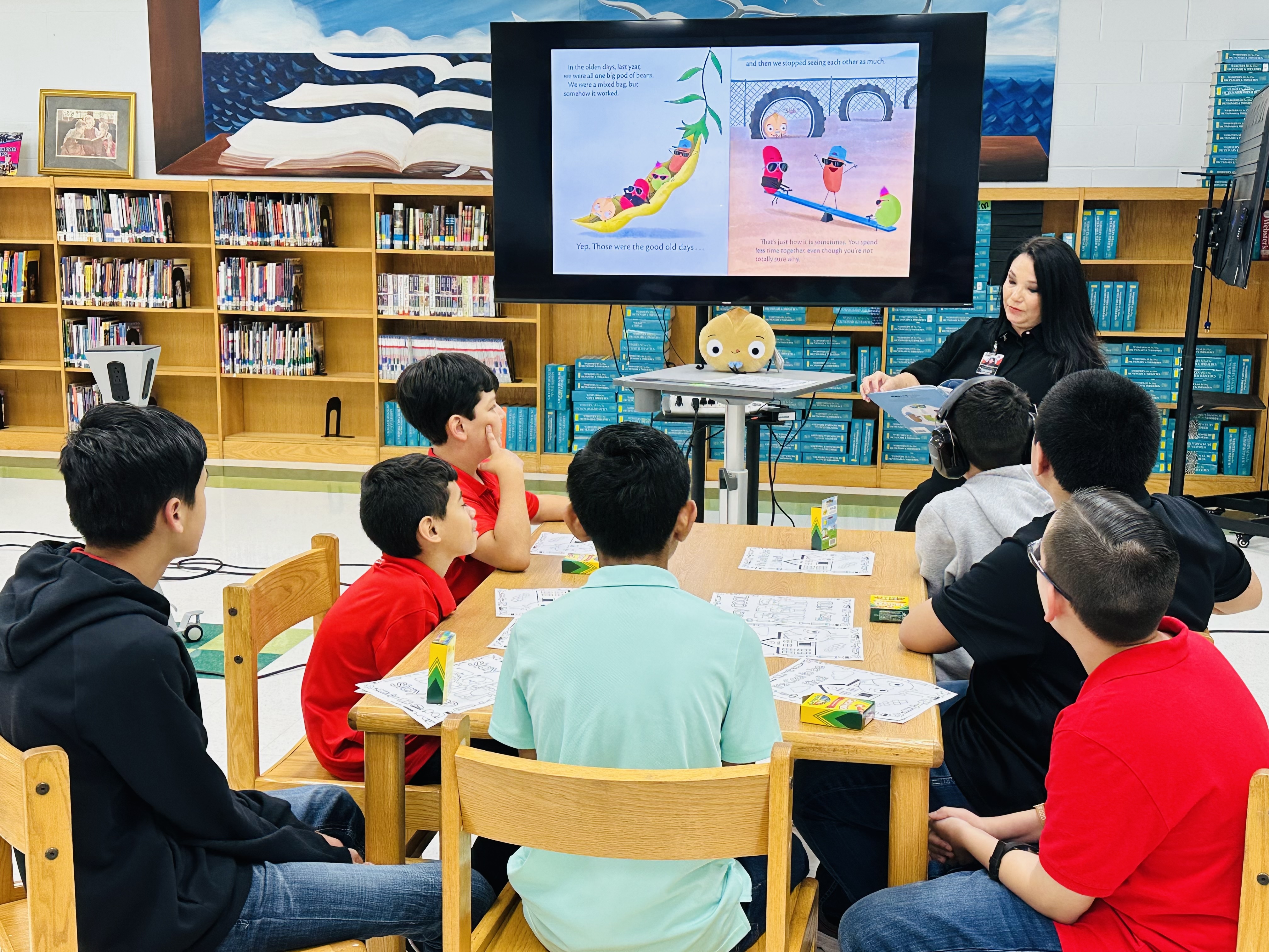 A teacher reads a story to a group of students seated at a table in a library.