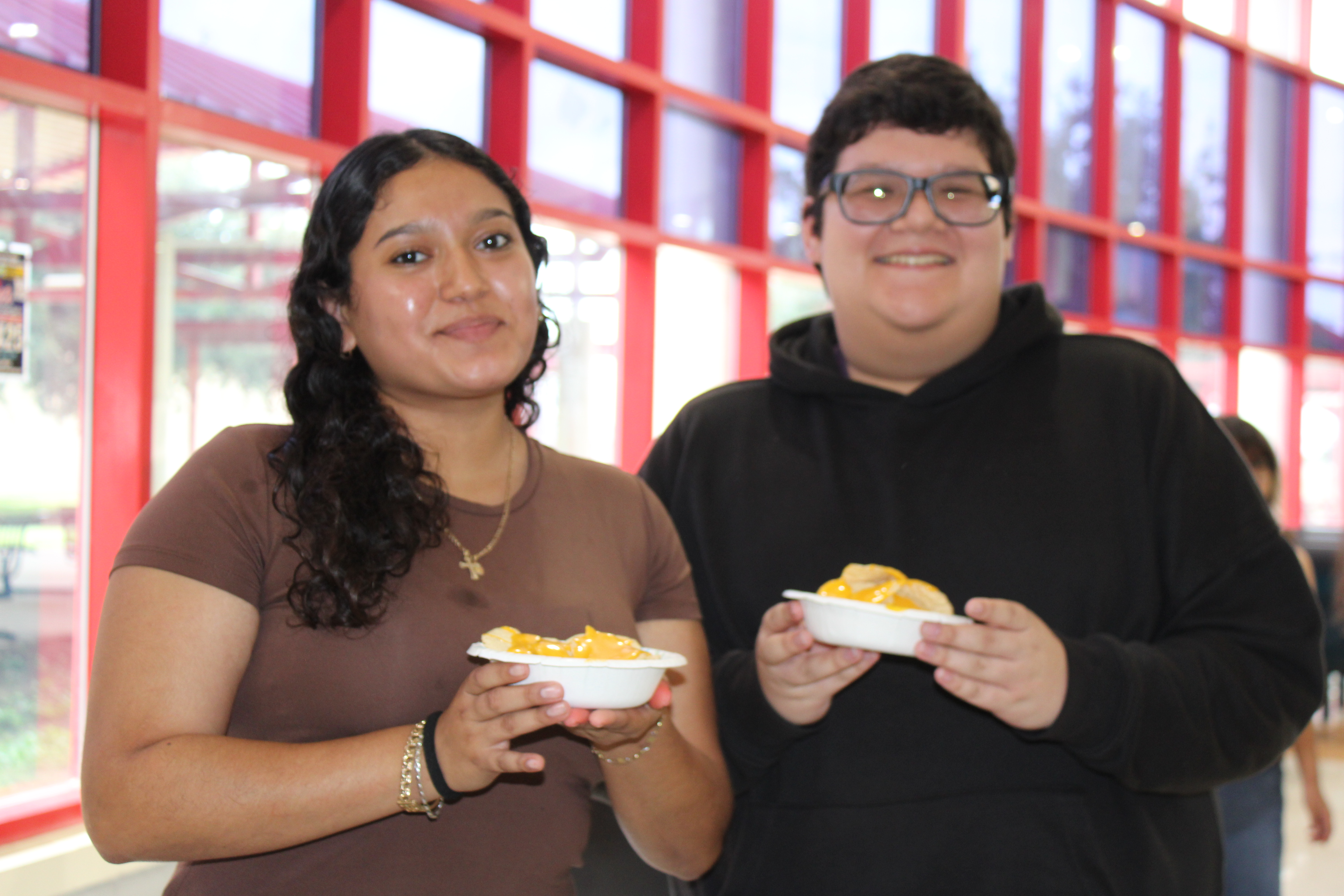 Two smiling people stand side-by-side, each holding a small white bowl with food inside.