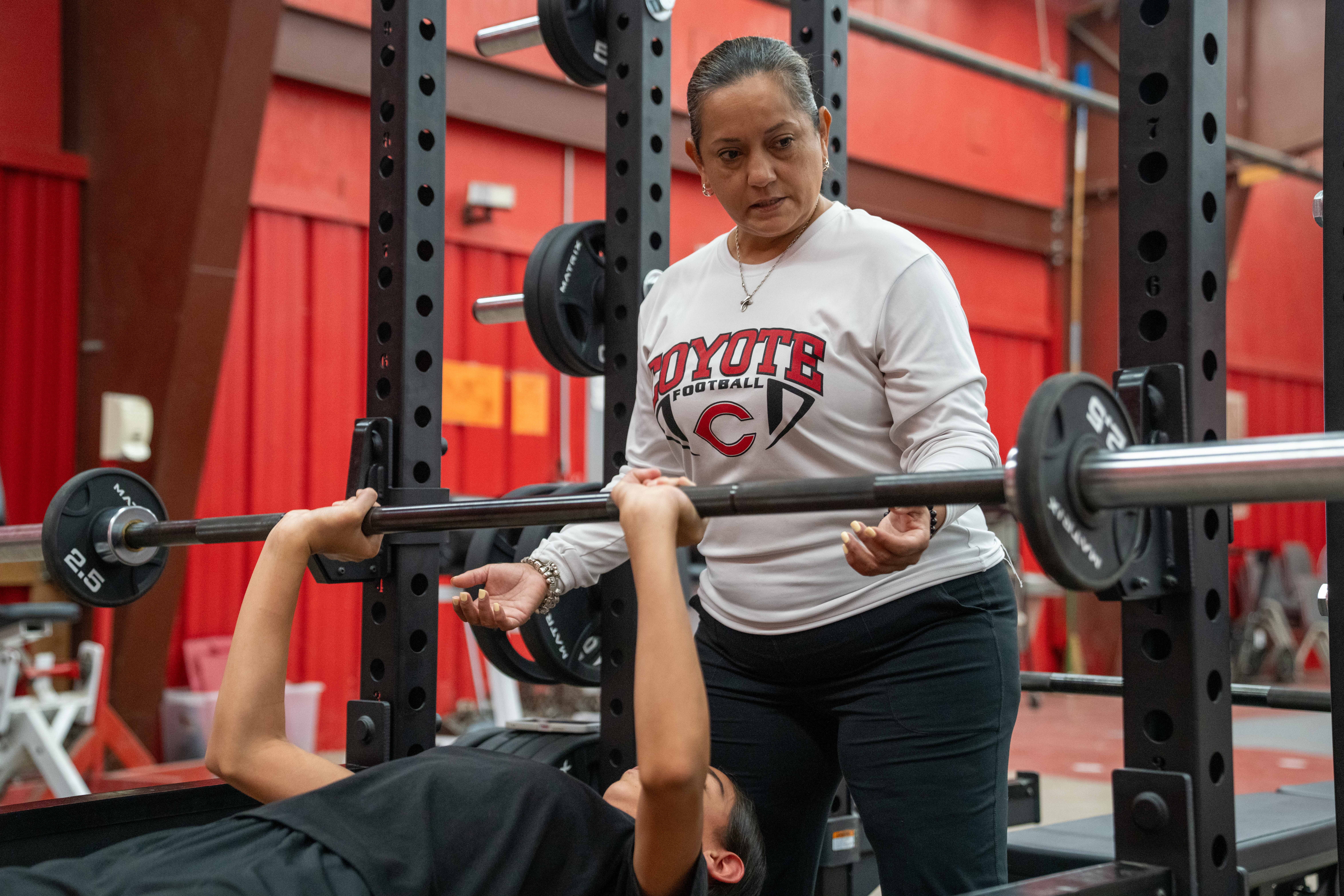 A coach spots a young athlete performing a bench press in a gym.
