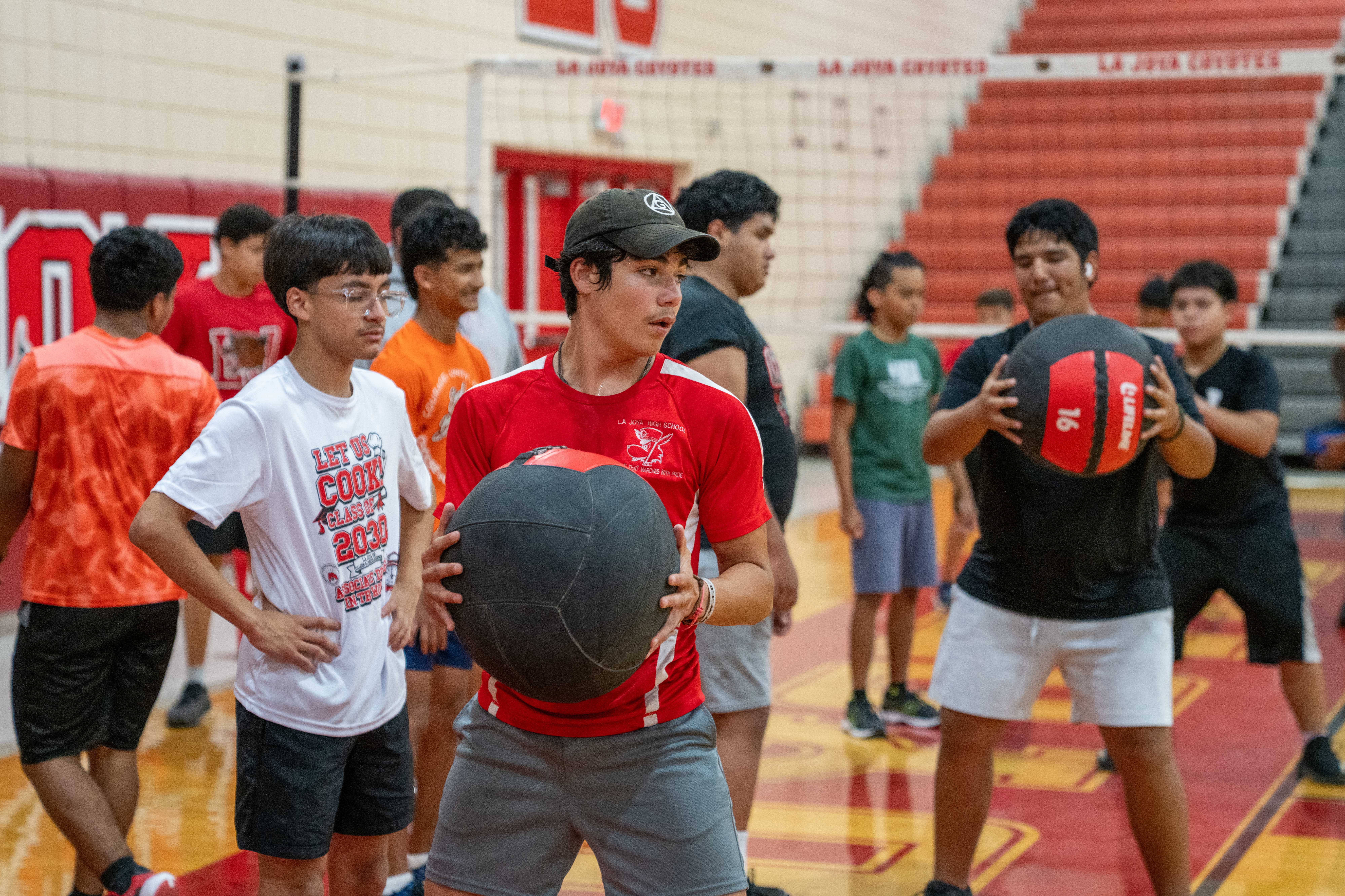 A group of young people in athletic wear stand on a polished gym floor, some holding medicine balls.