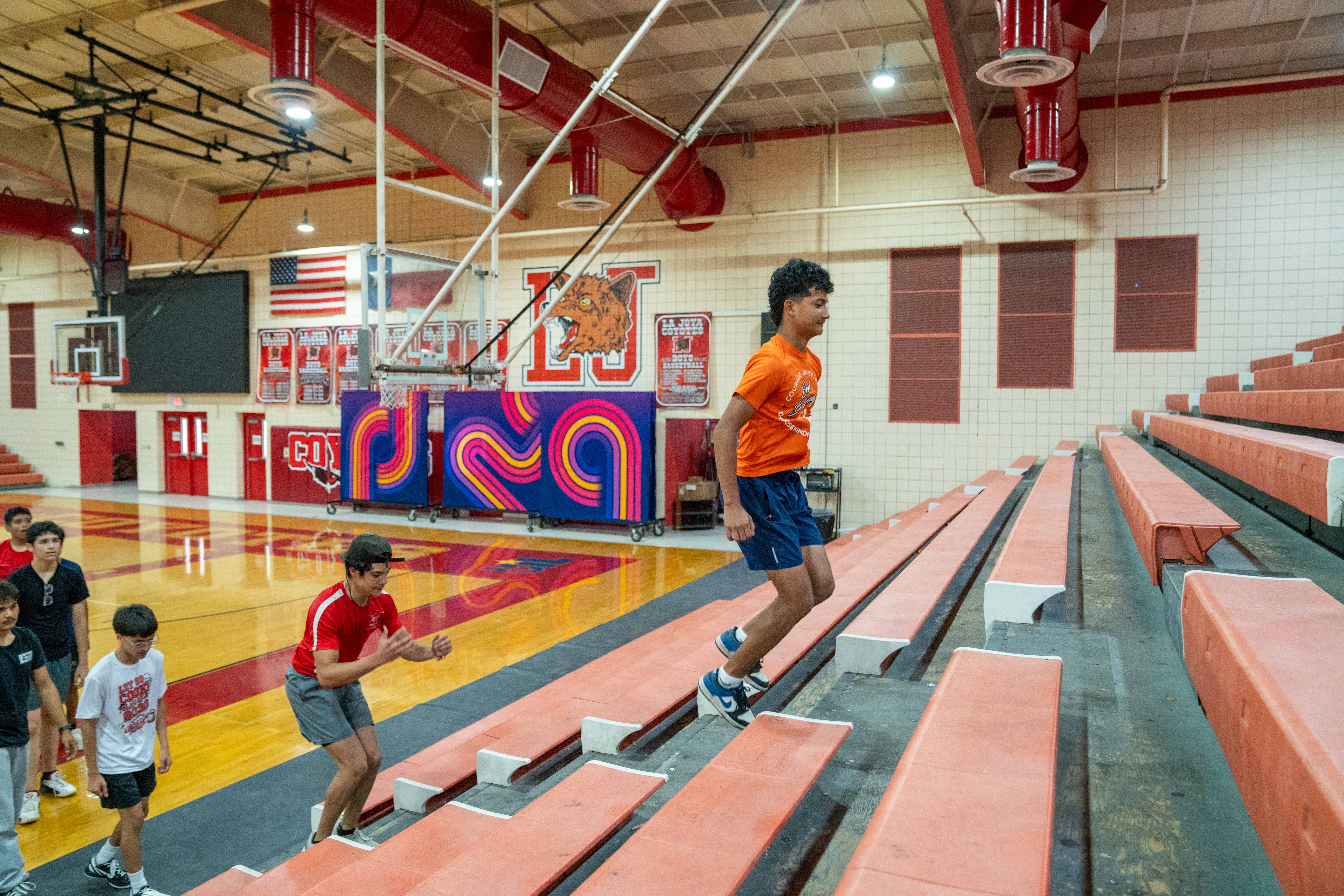 Teenagers practice basketball drills in a gymnasium, with one player running up bleachers.