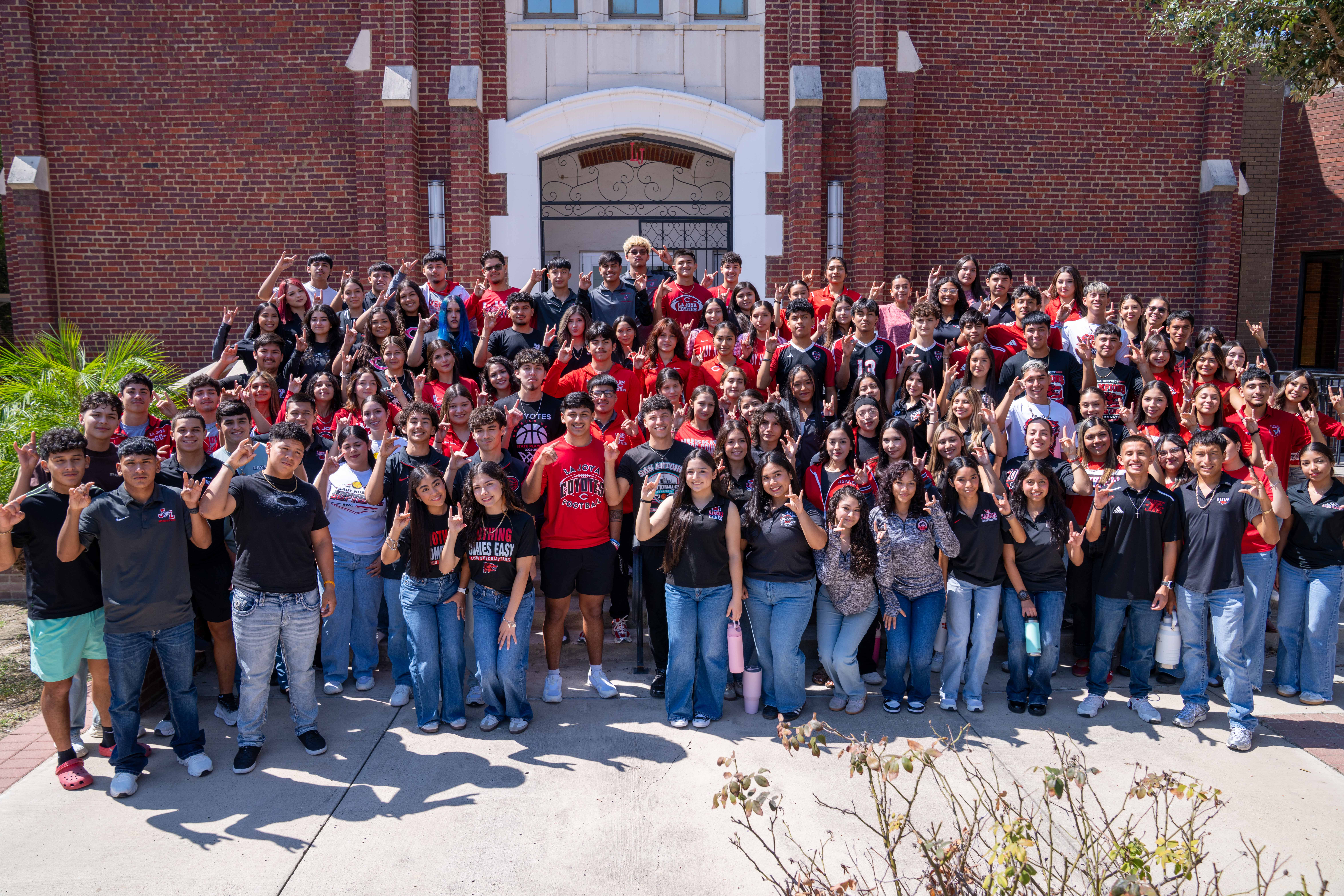 A large group of high school students in red and black athletic shirts pose outside a red brick building.