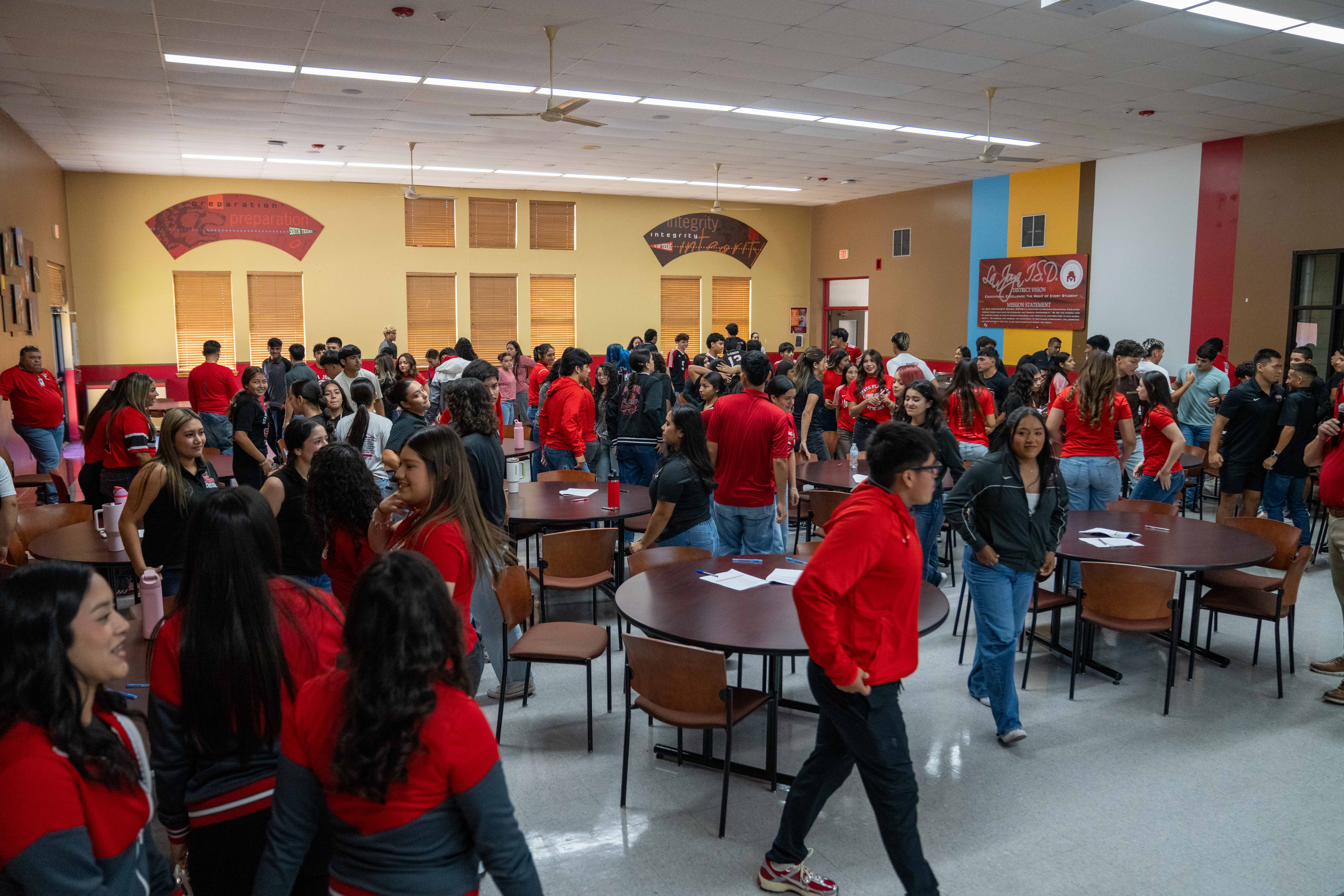 A large group of high school students gathers inside a brightly lit cafeteria with round tables and chairs.