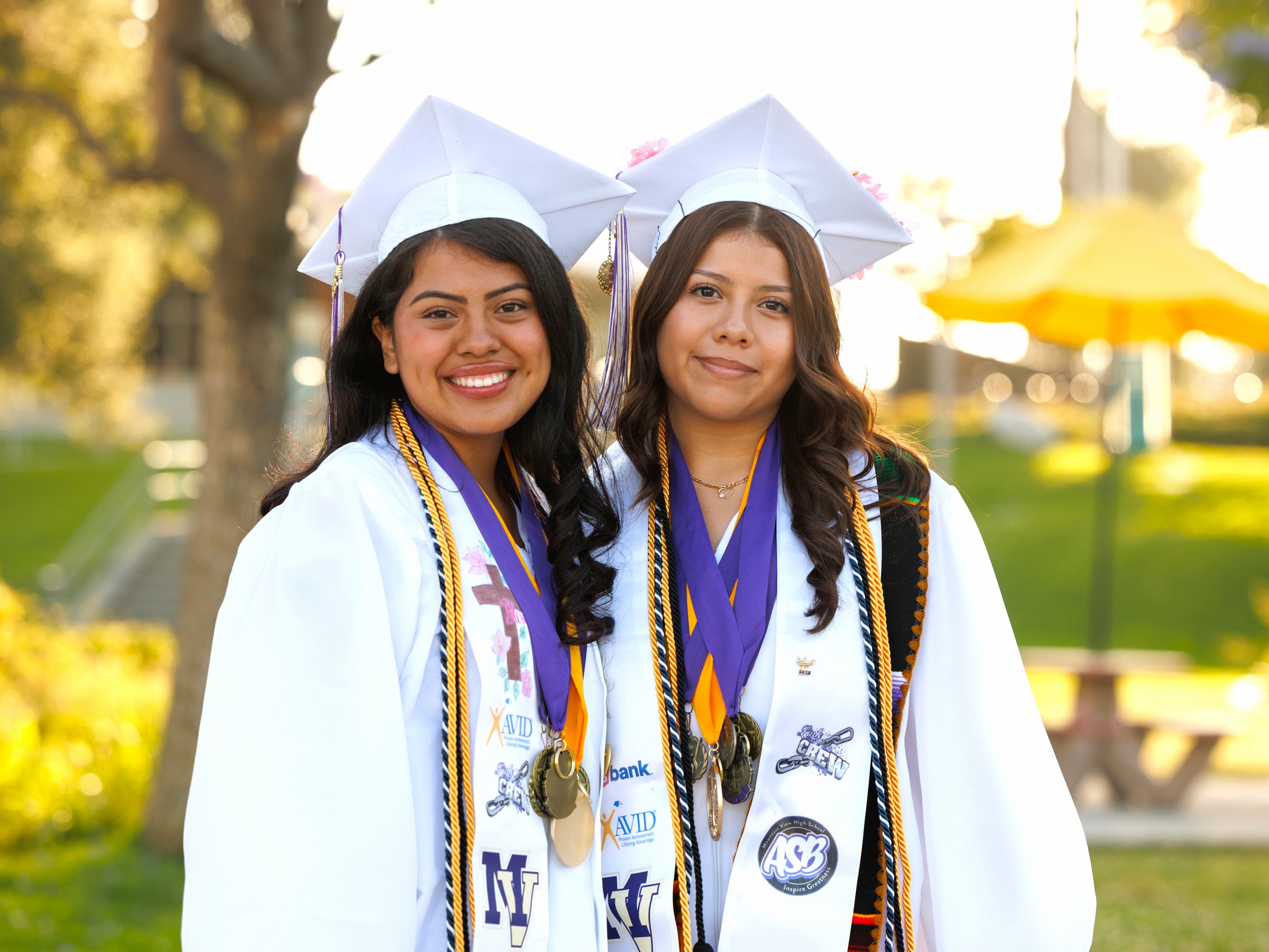 Two smiling graduates stand outdoors, wearing white caps and gowns adorned with purple stoles and medals.