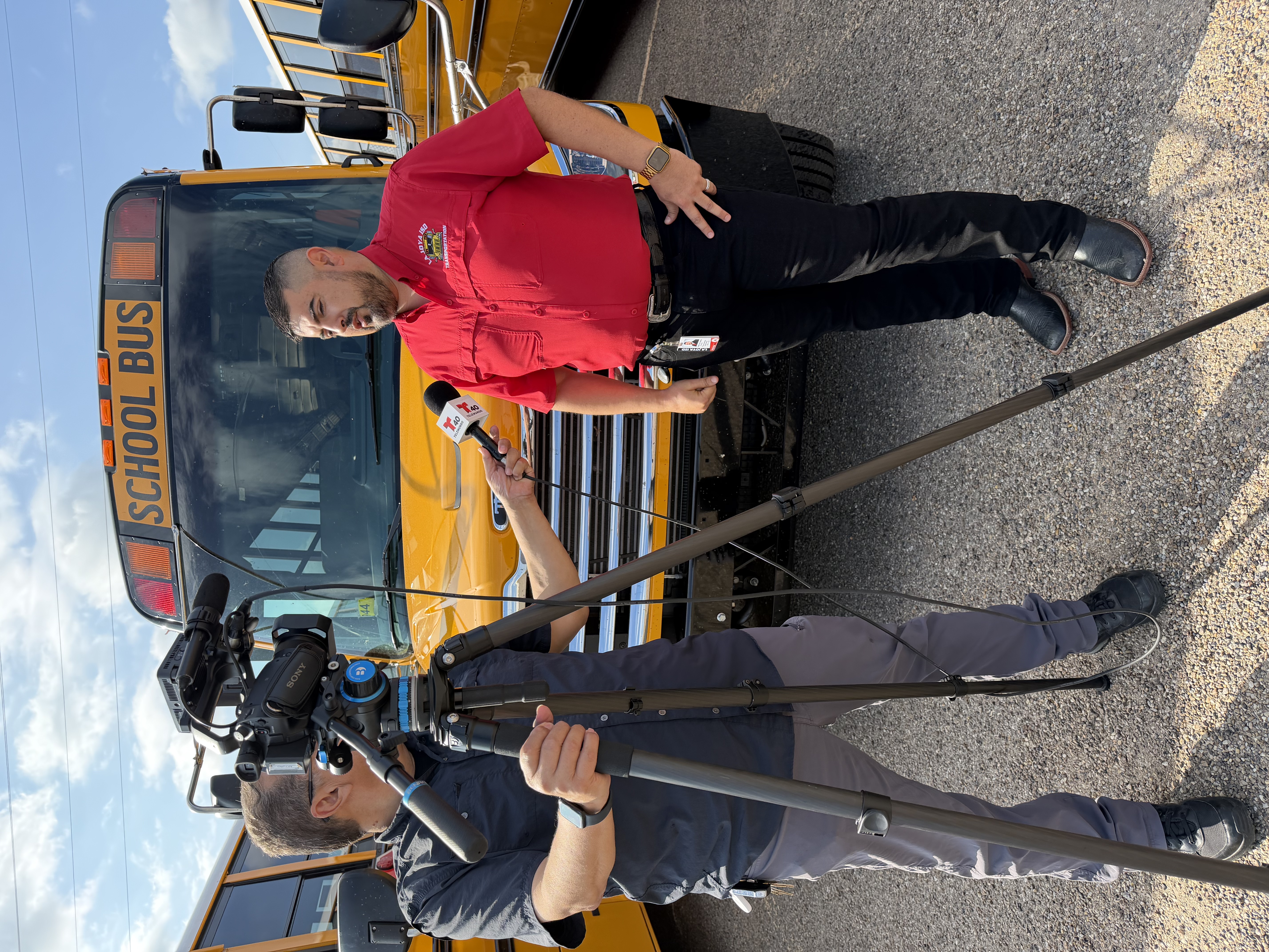 A news reporter wearing a red shirt and black pants stands in front of a yellow school bus, holding a microphone.