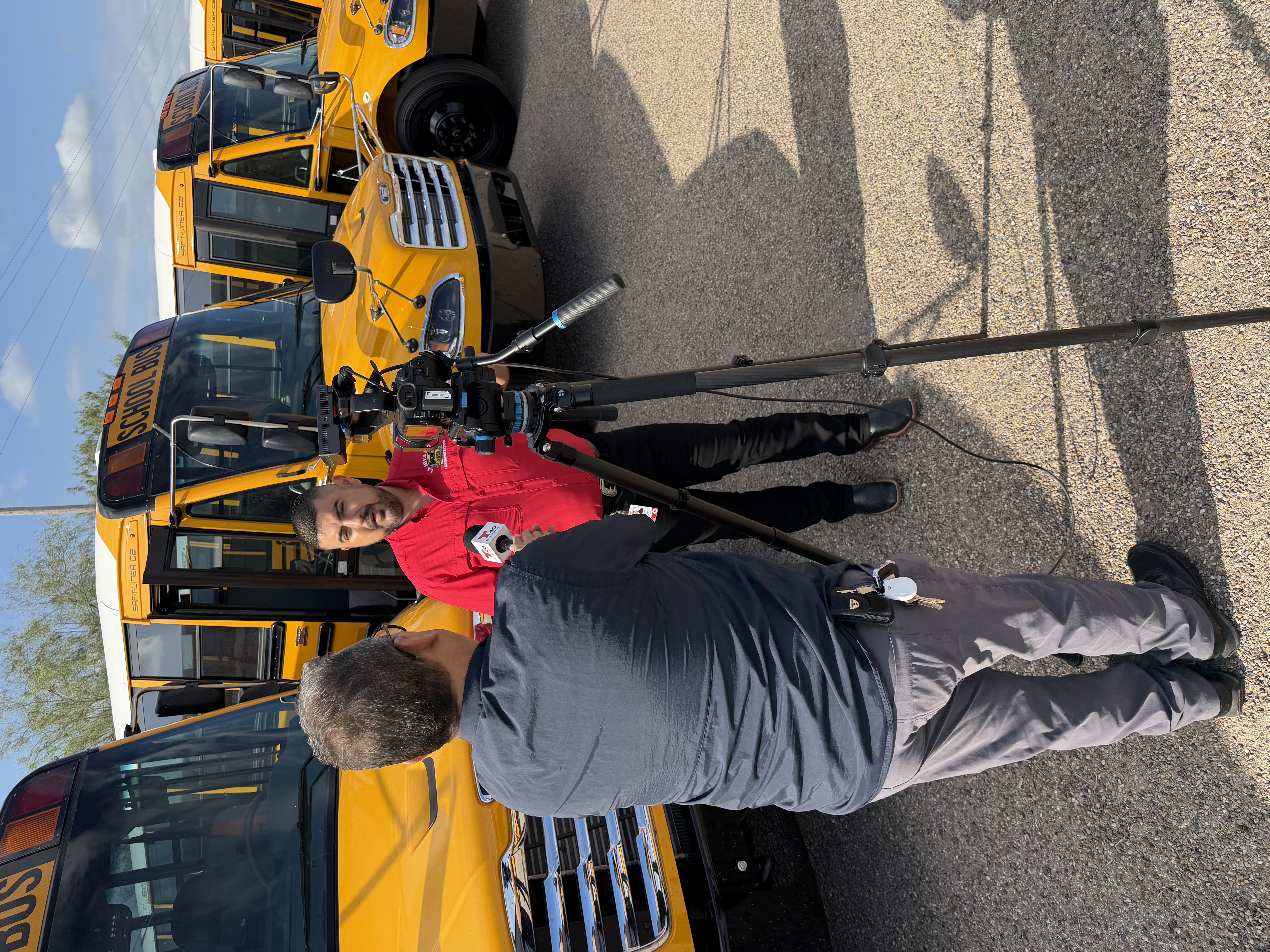 A reporter interviews a person in front of a row of parked yellow school buses.