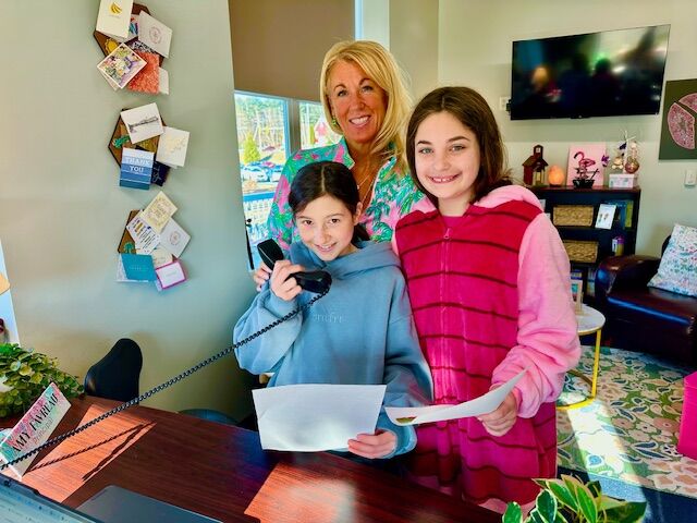 A woman smiles behind two young girls, one holding a phone and the other a paper, at a desk.