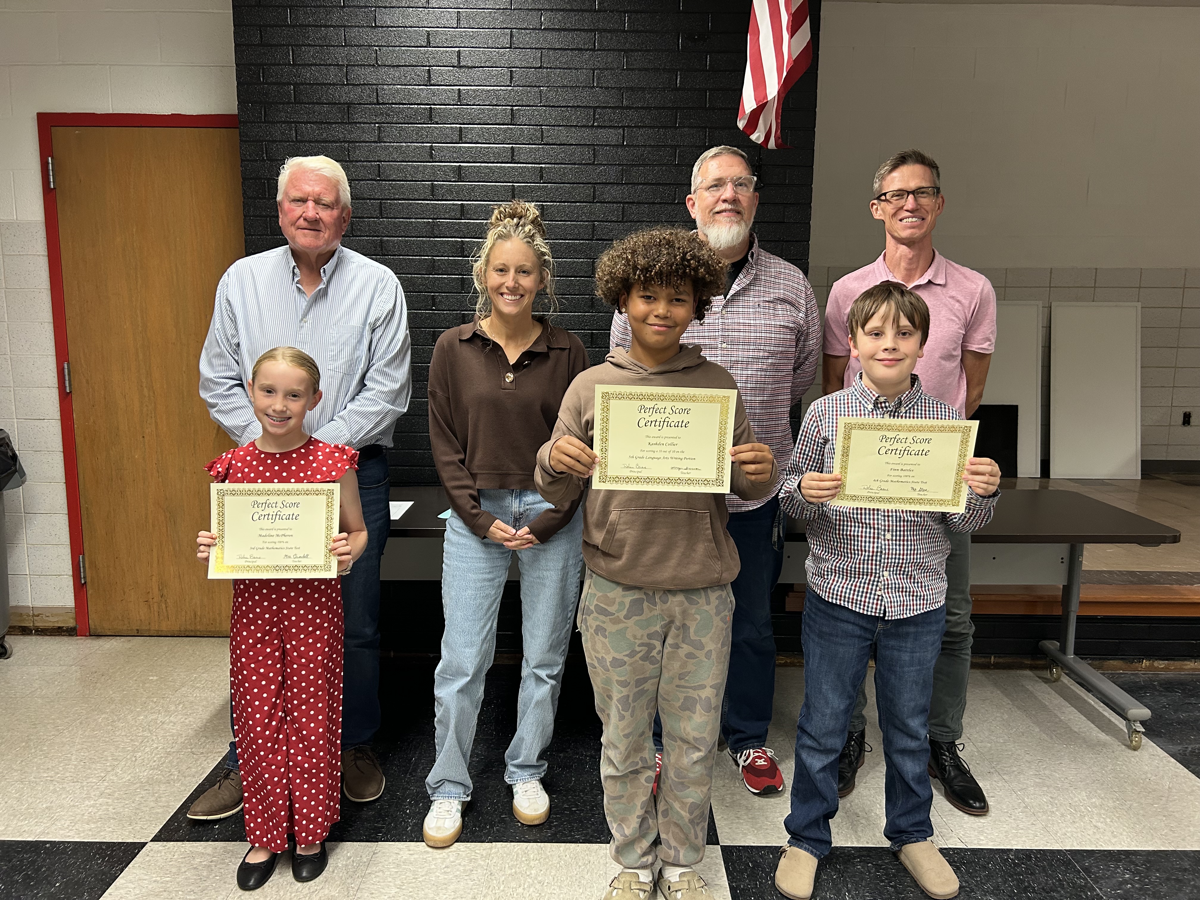 Three students hold up their Perfect Score certificates, smiling proudly alongside four adults in a school room.