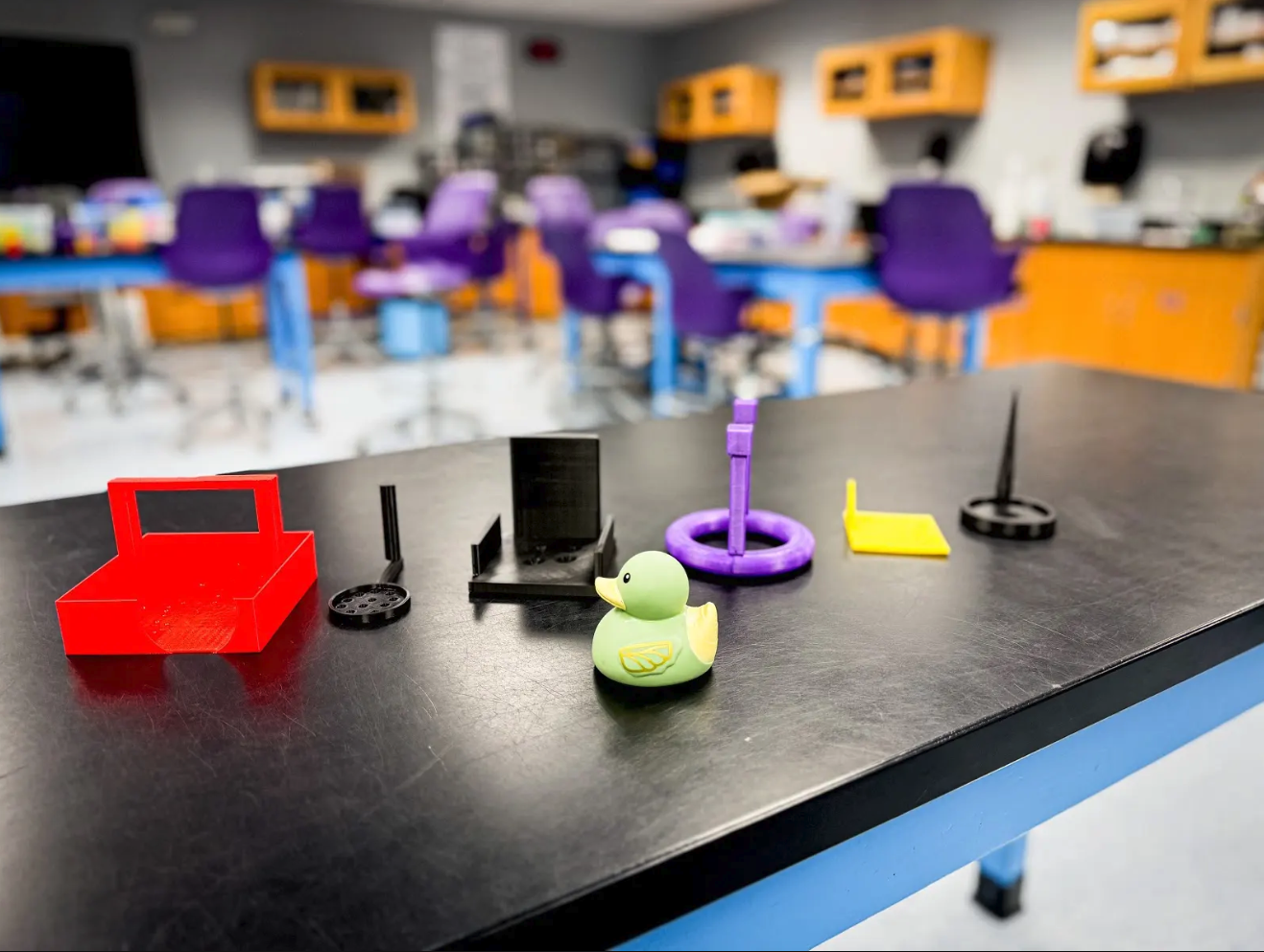 A collection of colorful 3D printed objects, including a duck, are displayed on a black table in a classroom.