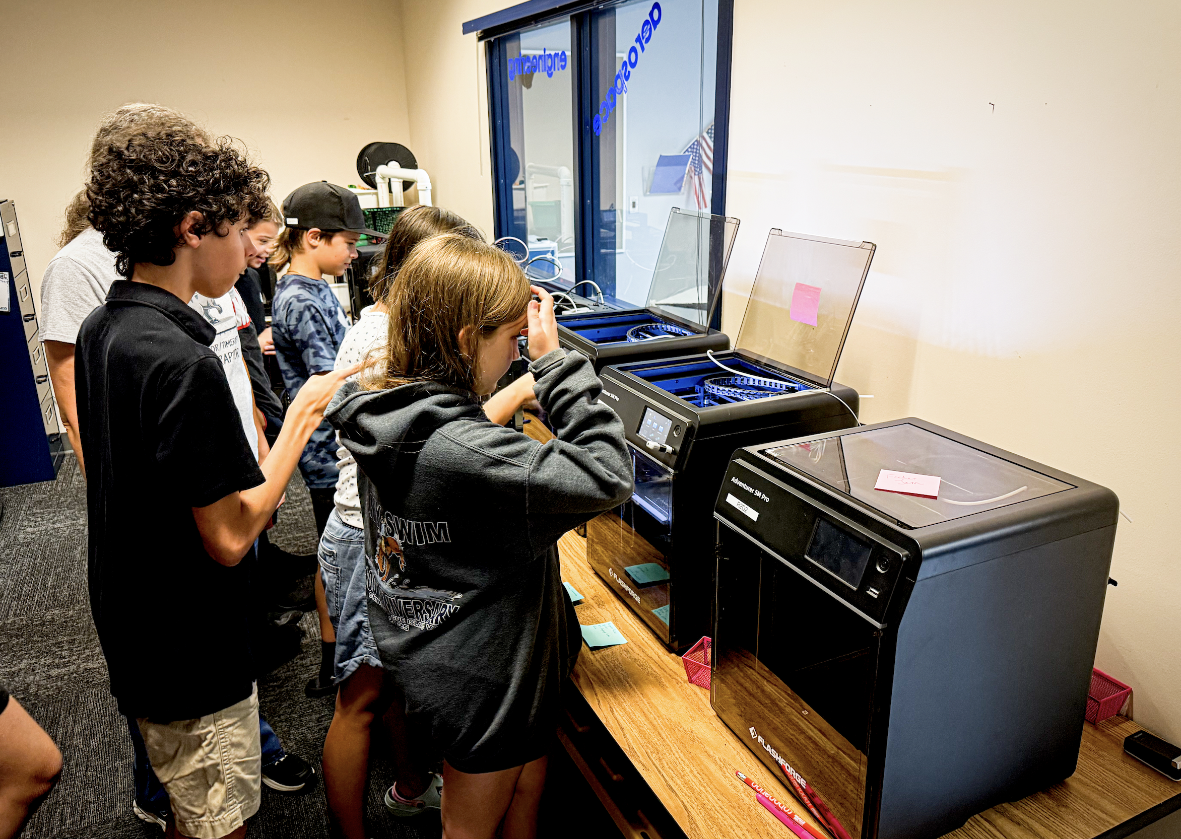 Children gather around 3D printers, observing the printing process.
