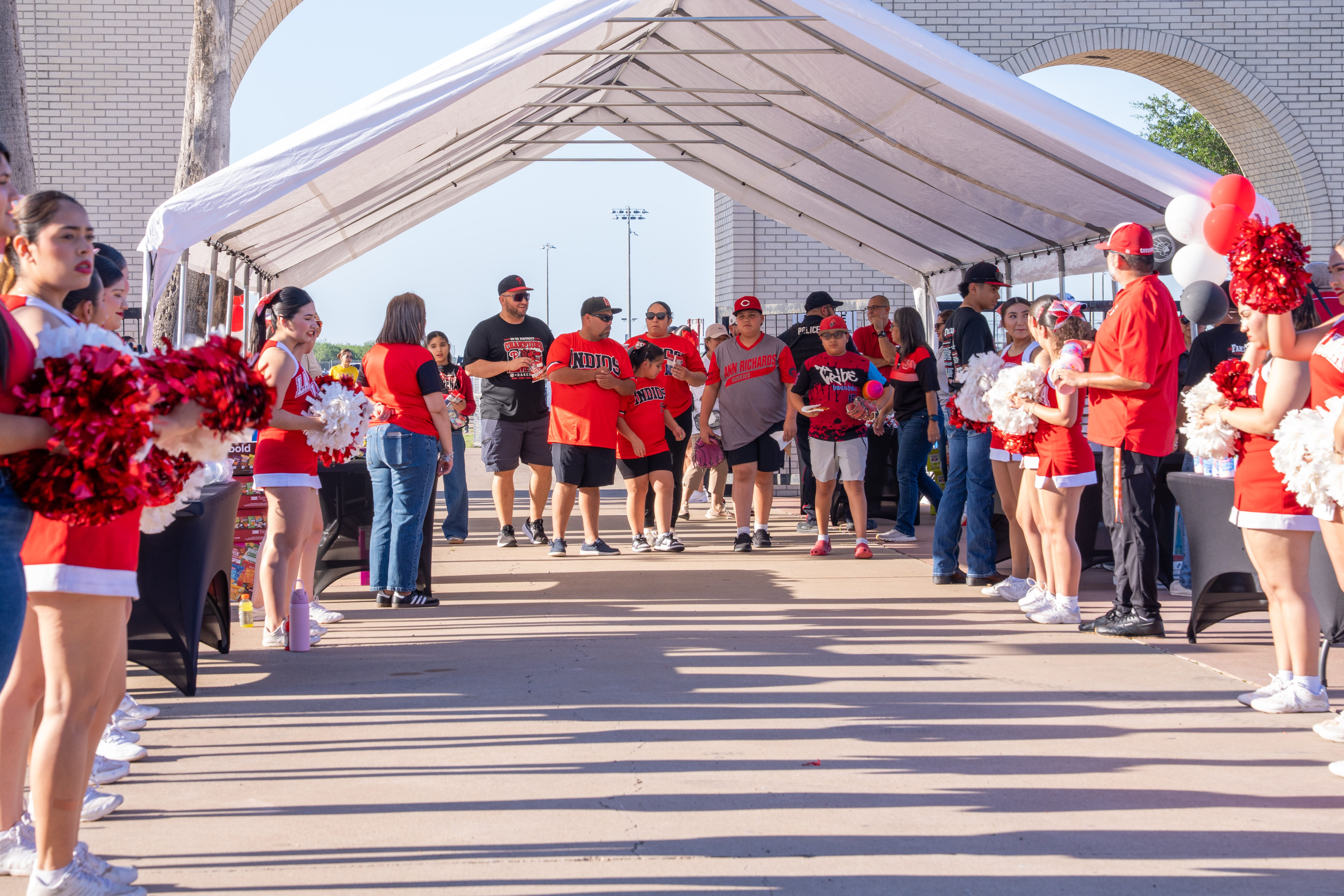 A group of people in red and black attire, some holding pom-poms, stand under a white tent.