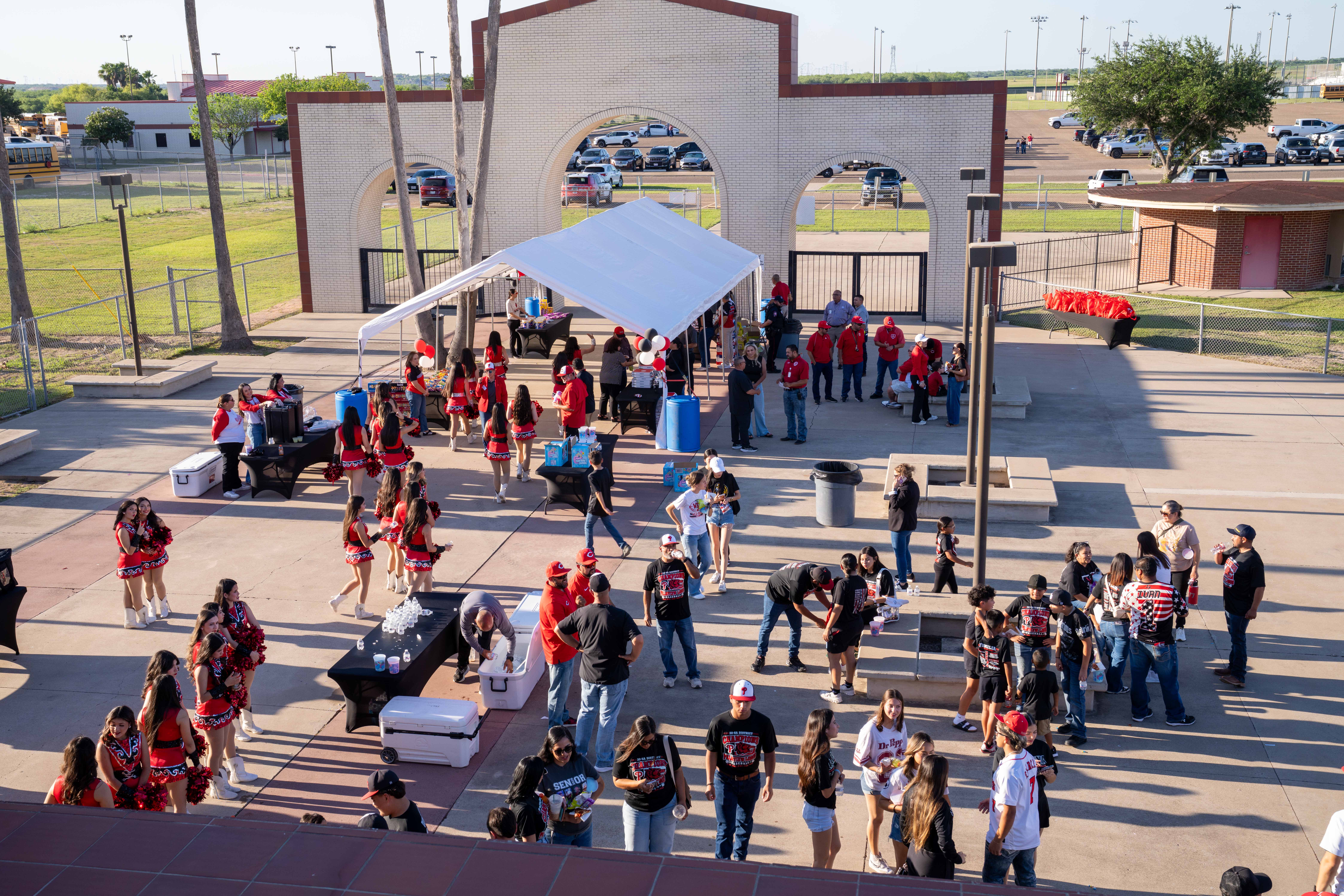 A group of people, some in cheerleading uniforms, gather under a white tent at an outdoor event.