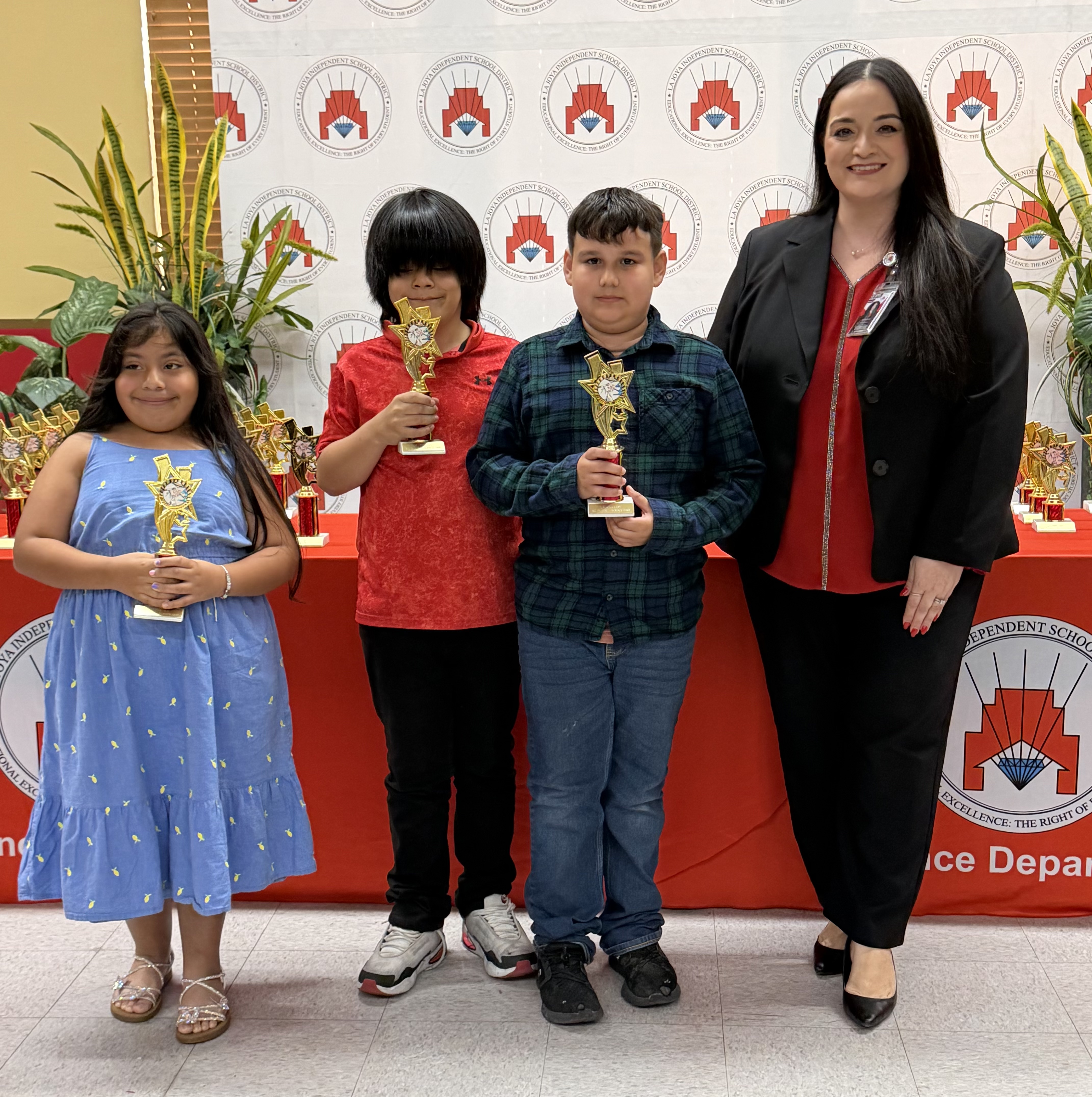 A woman in a black blazer stands with three children, each holding a trophy, in front of a red backdrop with school district logos.