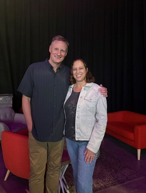 A smiling man and woman stand together in front of a black curtain, with red chairs and a sofa visible in the background.