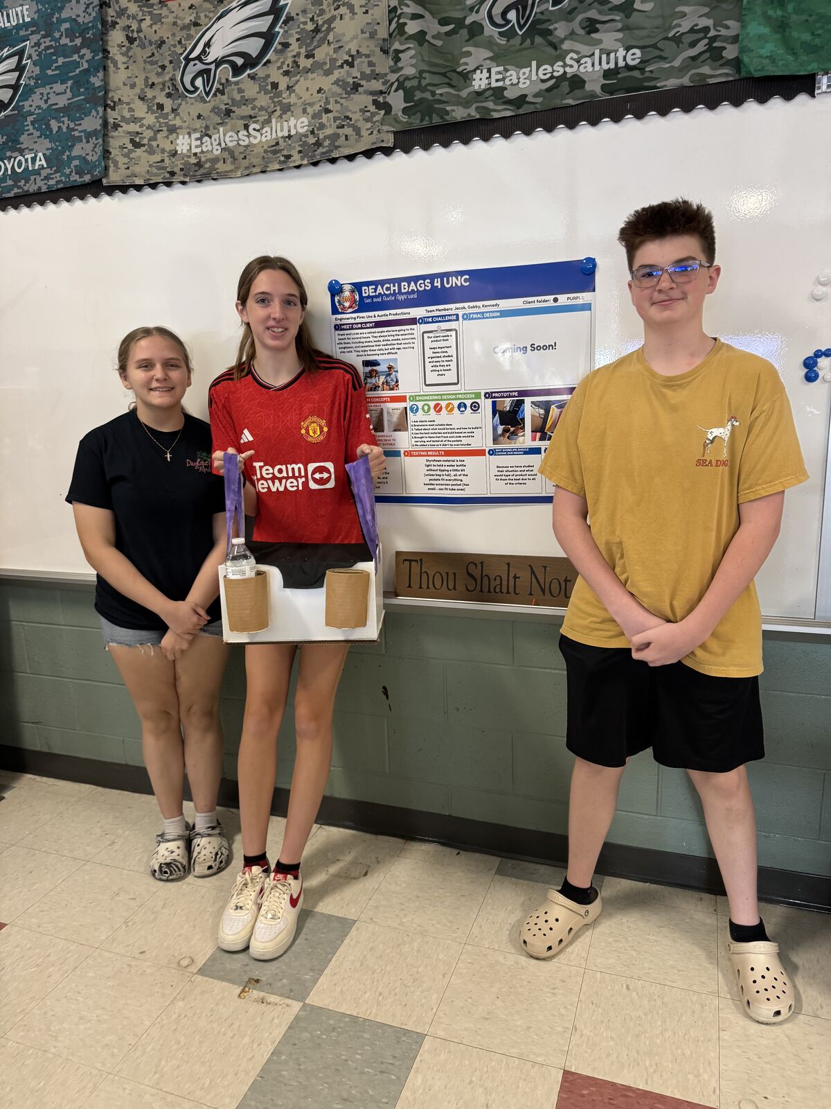 Three students stand in front of a presentation board titled 'BEACH BAGS 4 UNC', showcasing a prototype.