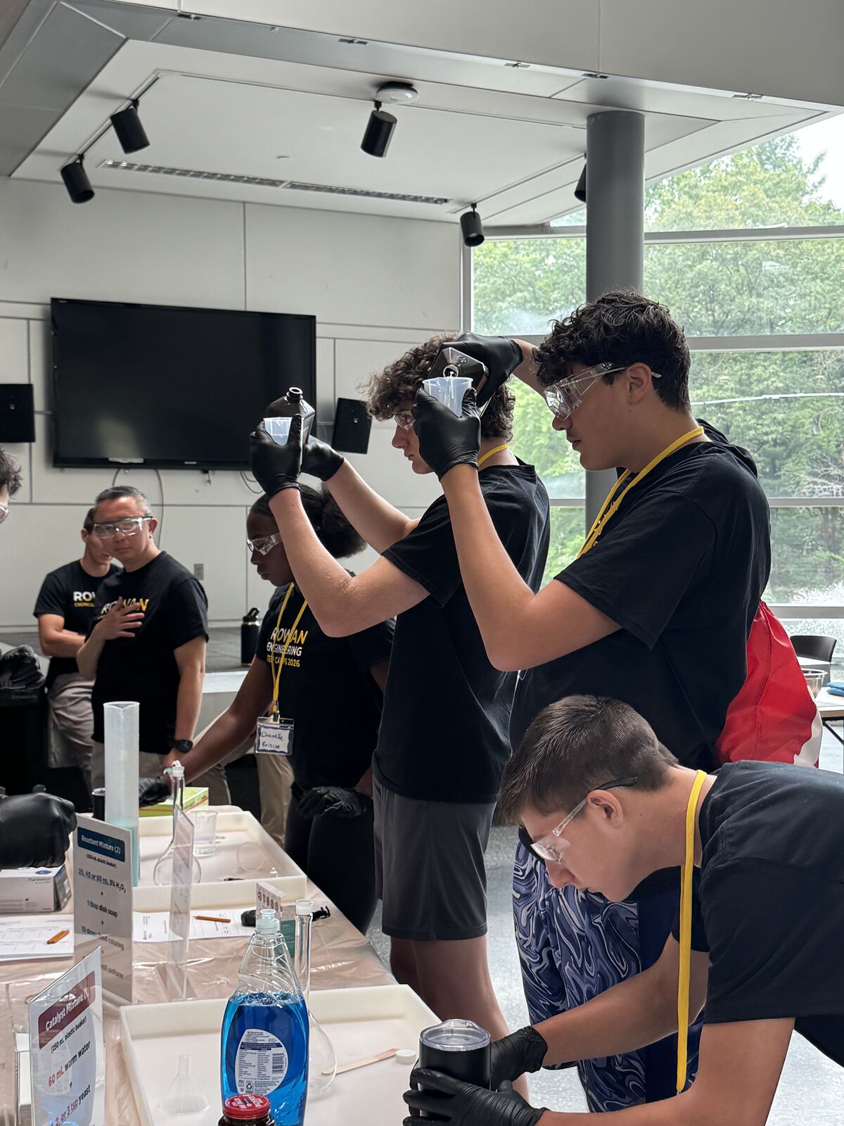 Students in black shirts and safety goggles conduct a science experiment, pouring liquids from beakers.